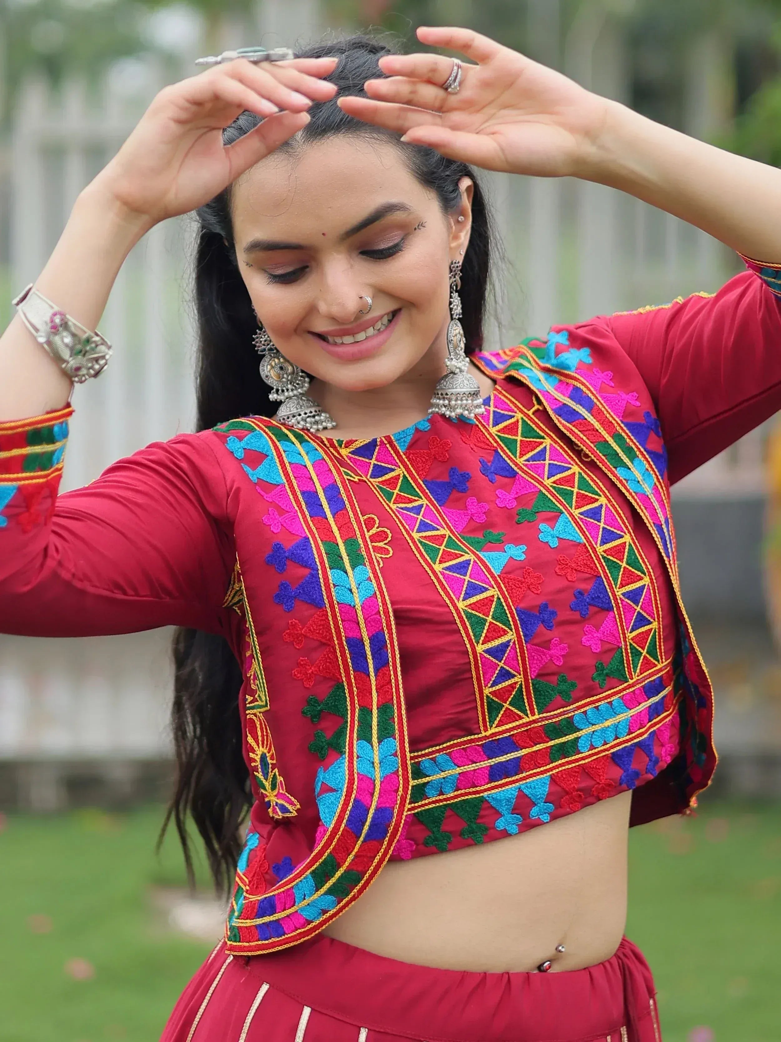 Woman in vibrant red ethnic choli with colorful embroidery, paired with traditional jewelry and nose ring, outdoors.