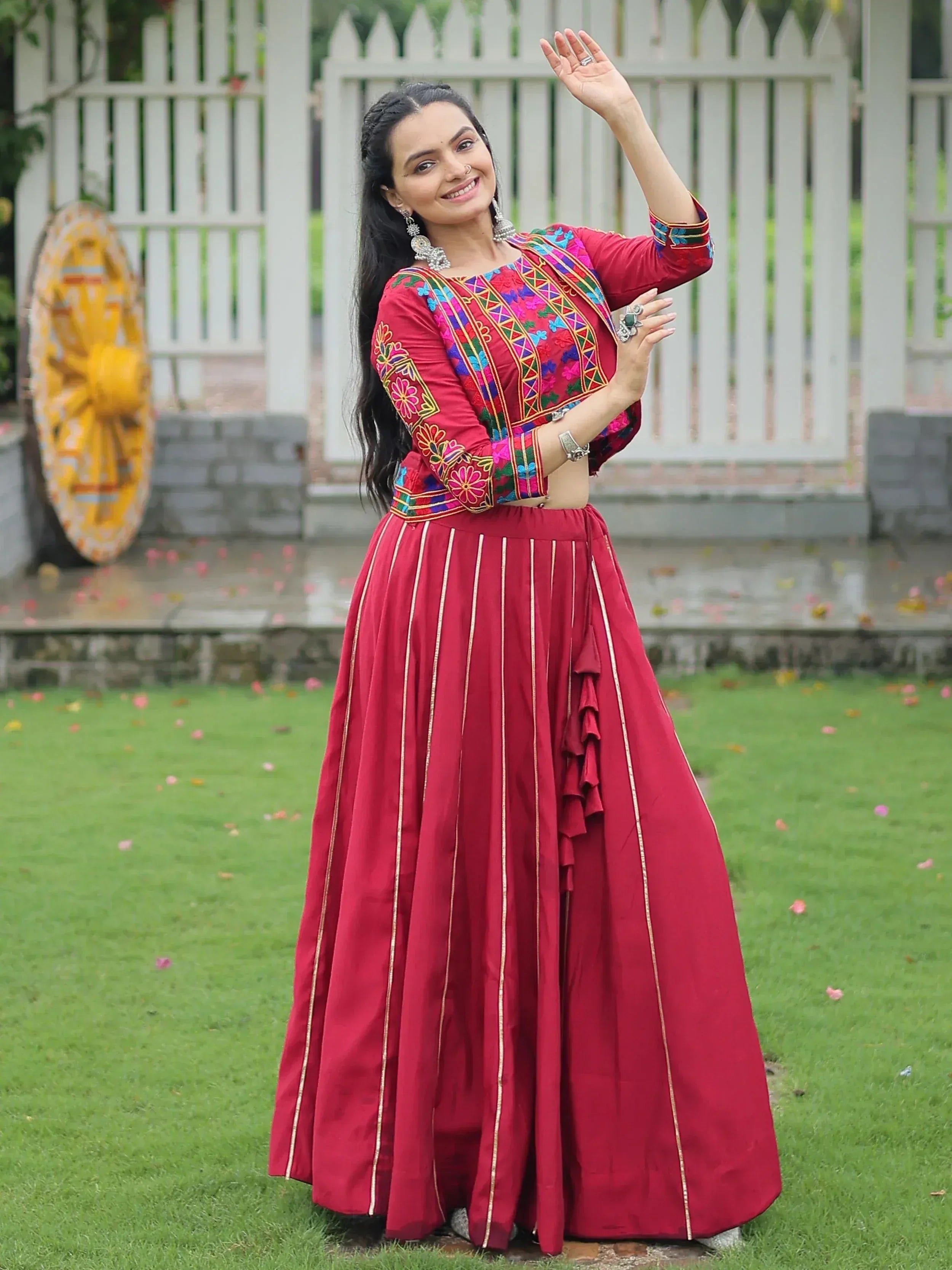 Woman in red embroidered lehenga choli with floral patterns, standing outdoors on grass near white gate