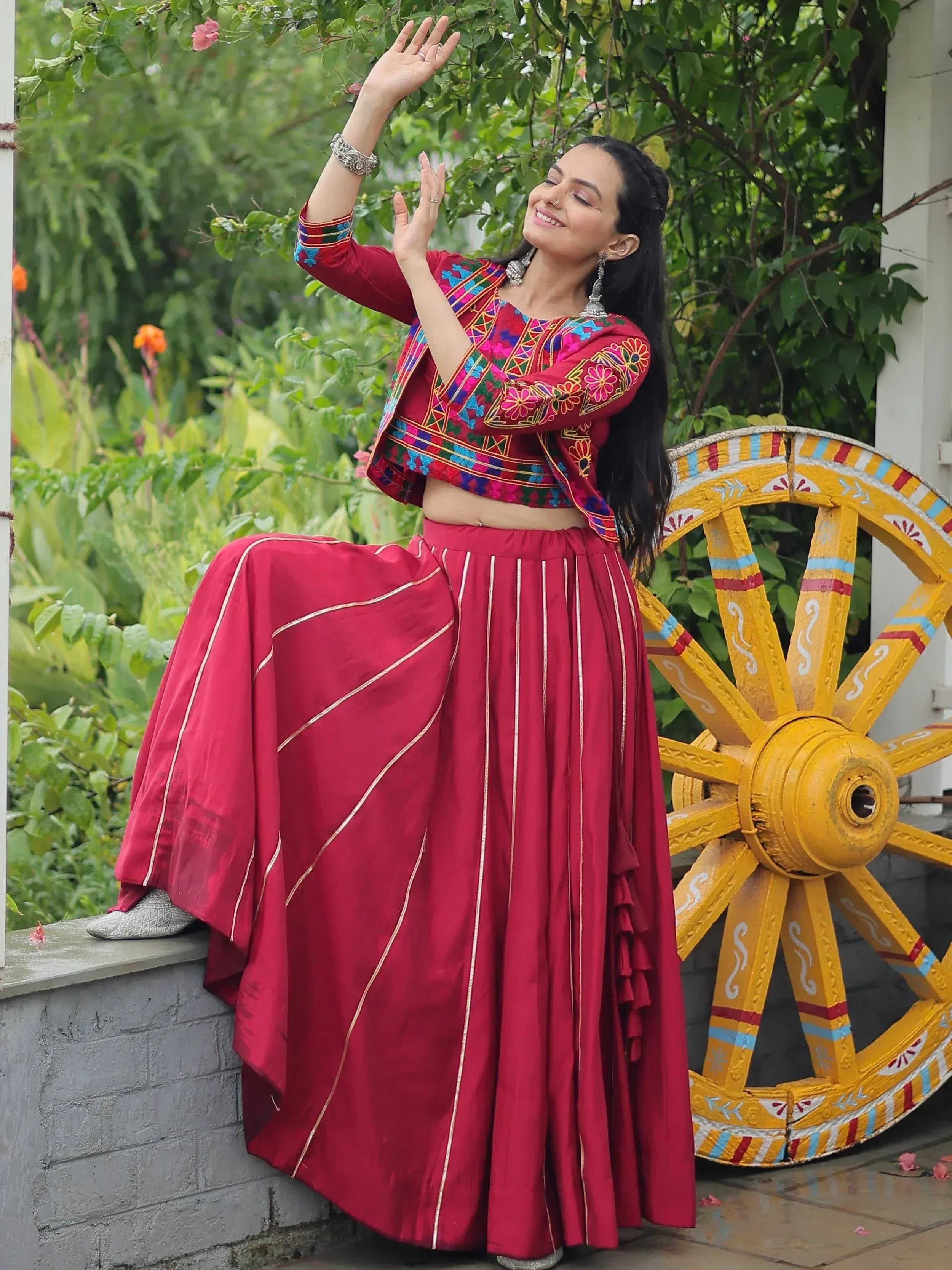 Woman in red embroidered lehenga choli with gold stripes, posing near a colorful wheel in a garden