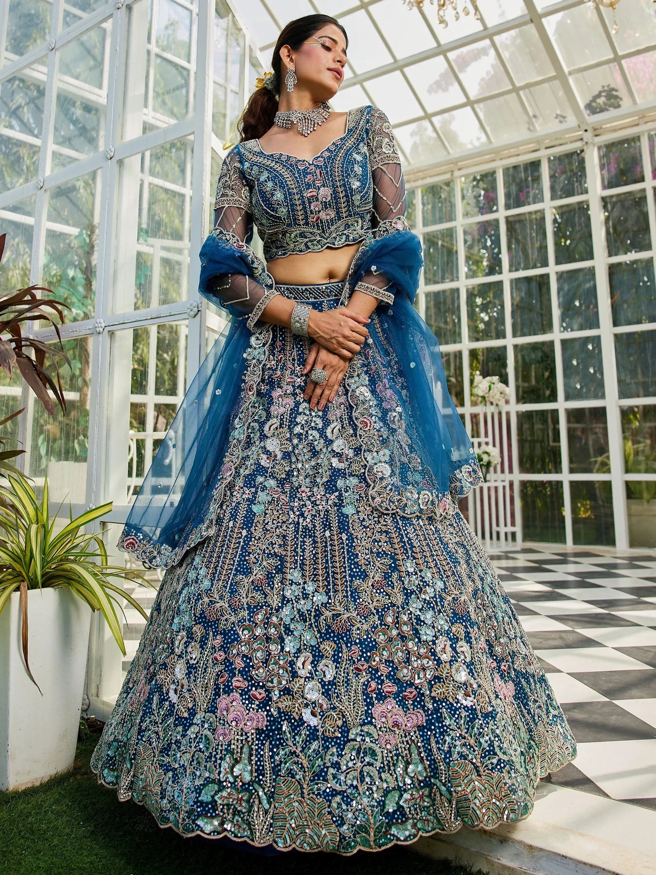 Blue embroidered lehenga choli with floral motifs, sheer sleeves, and dupatta, styled in a glass conservatory.