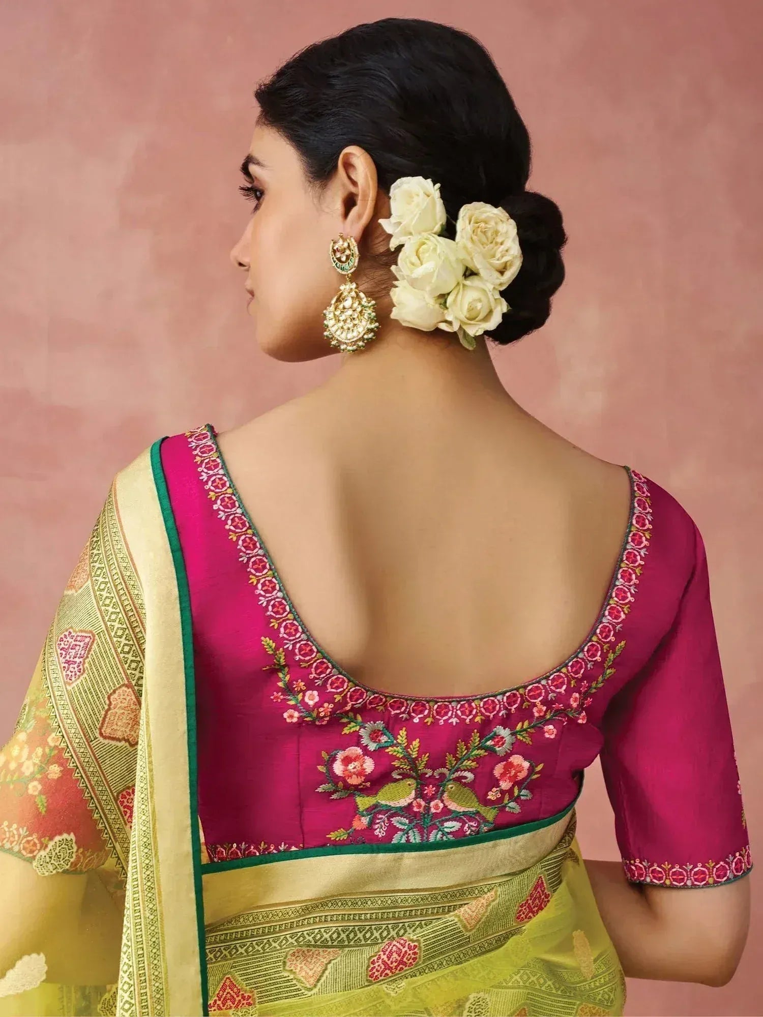 Woman in pink embroidered saree blouse with floral back design, gold earrings, and white rose hair accessory