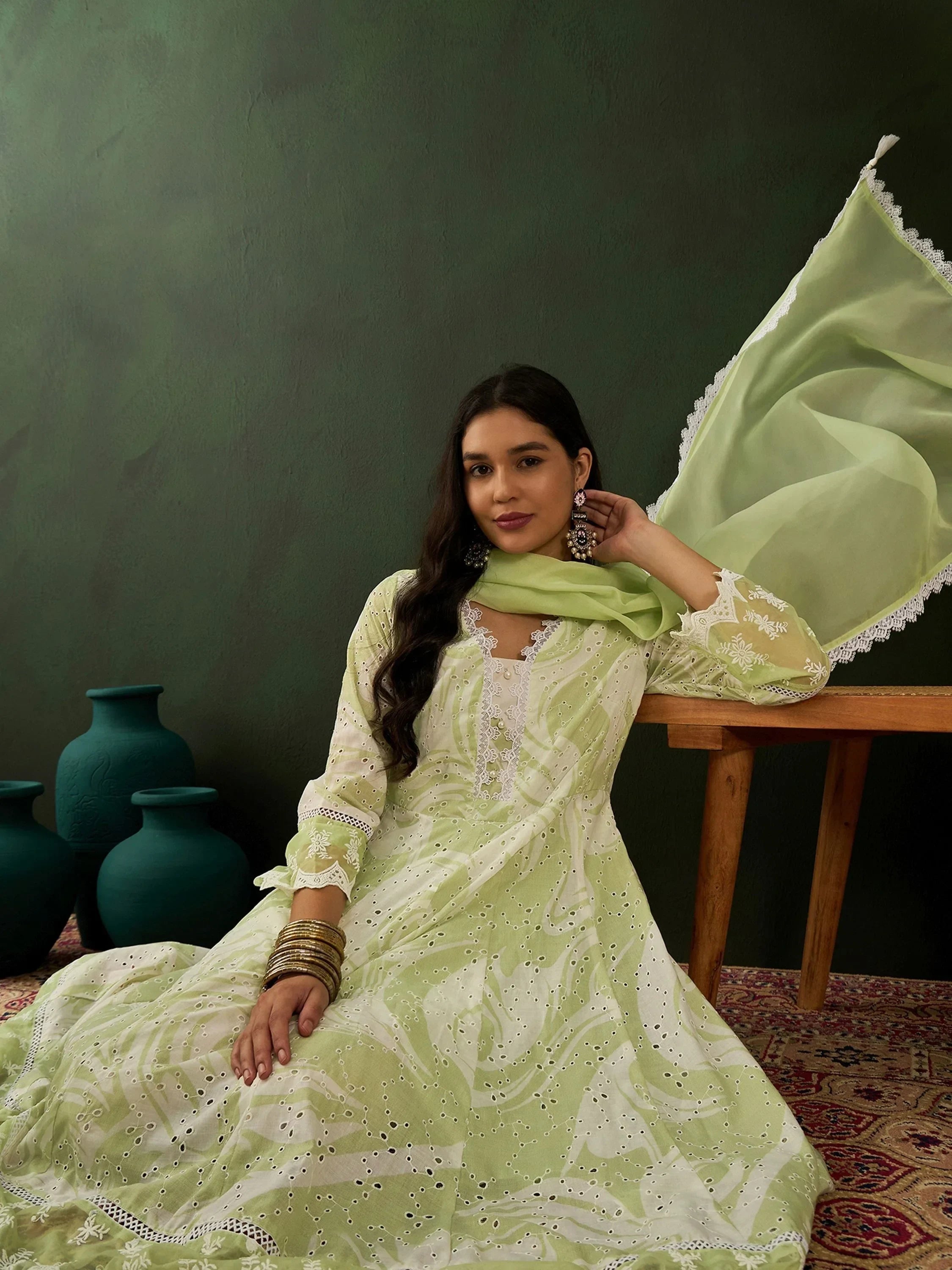 Woman in light green embroidered ethnic gown with dupatta, sitting on patterned rug