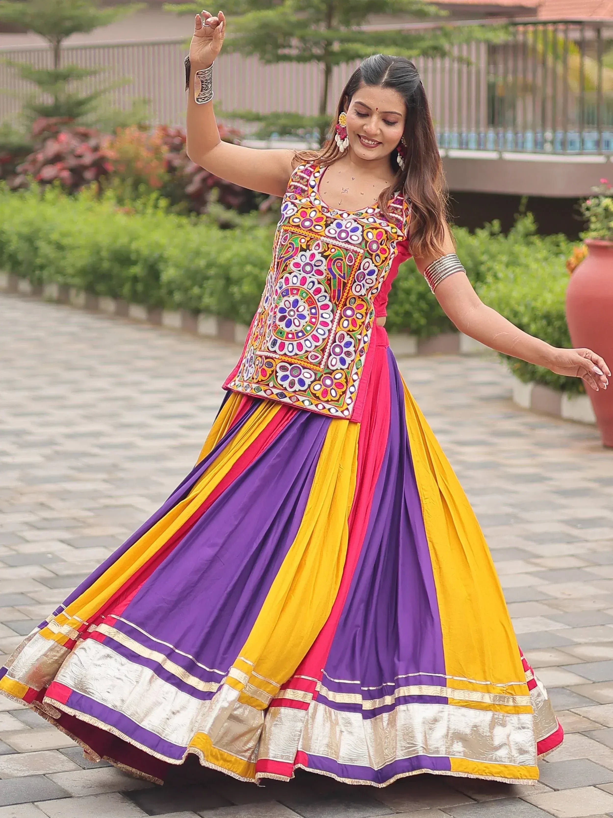 Woman in colorful embroidered ethnic lehenga choli with vibrant purple, yellow, and pink pleated skirt, dancing outdoors.