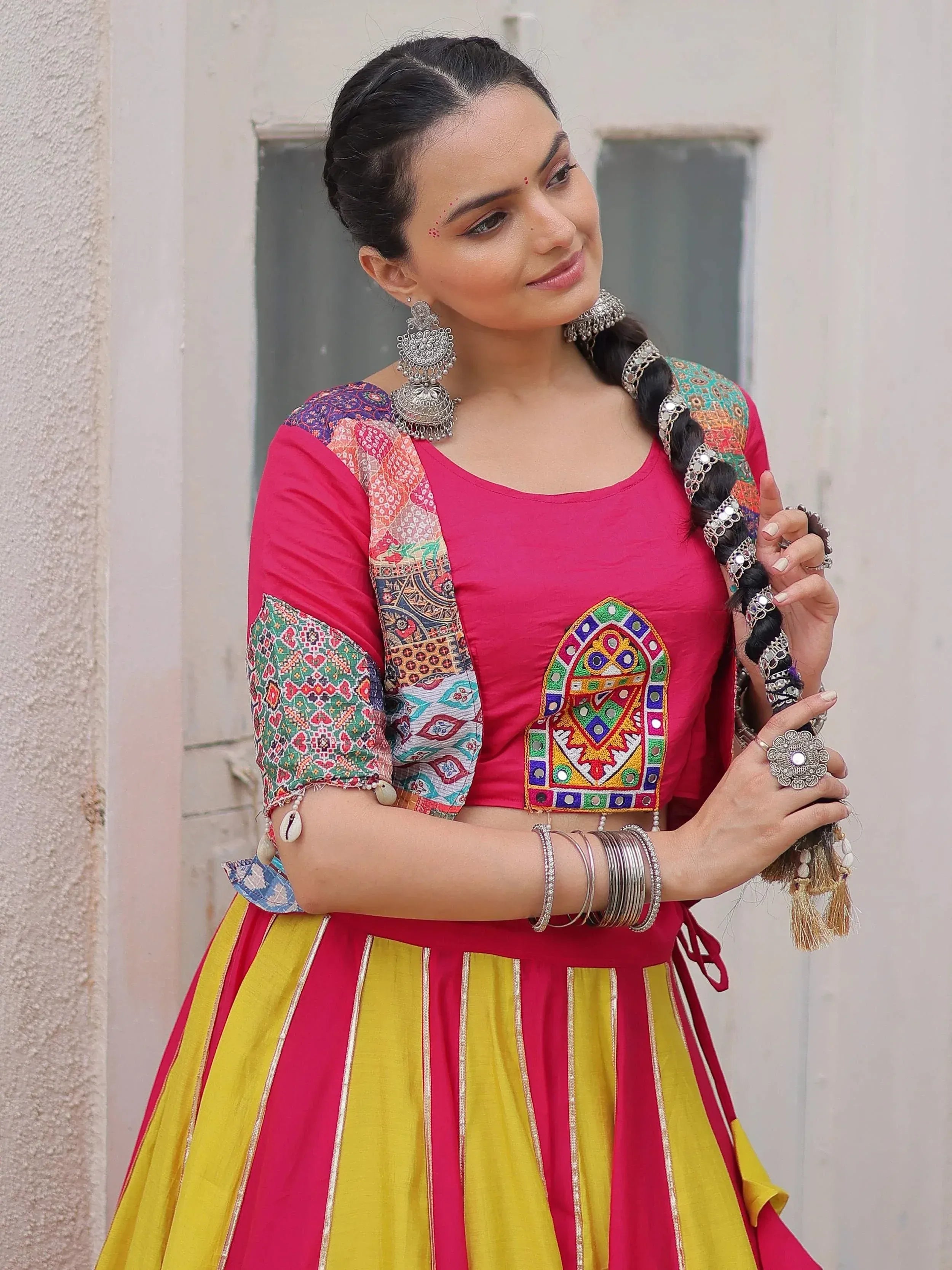 Woman in vibrant pink and yellow ethnic lehenga choli with mirror work, traditional jewelry, and braided hair.