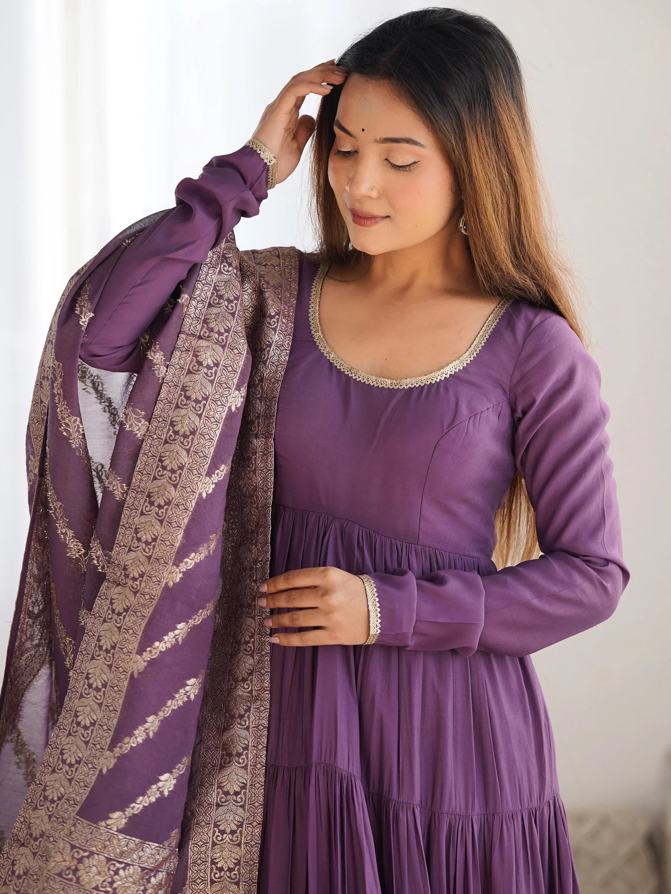 Woman in a purple Anarkali suit with gold embroidered dupatta, posing indoors