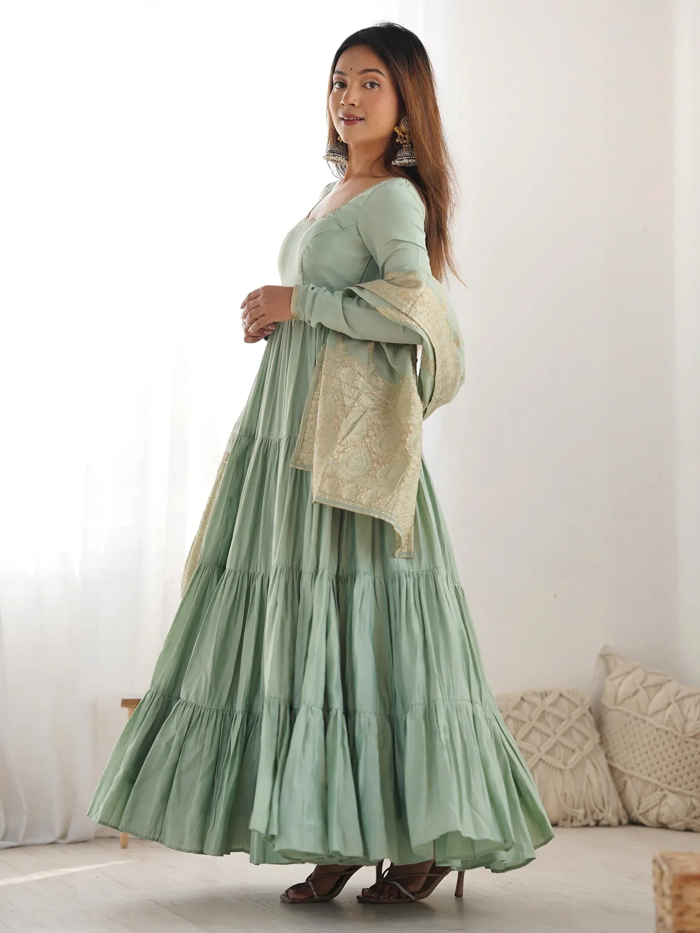 Woman in pastel green ethnic gown with gold dupatta and statement earrings, standing indoors