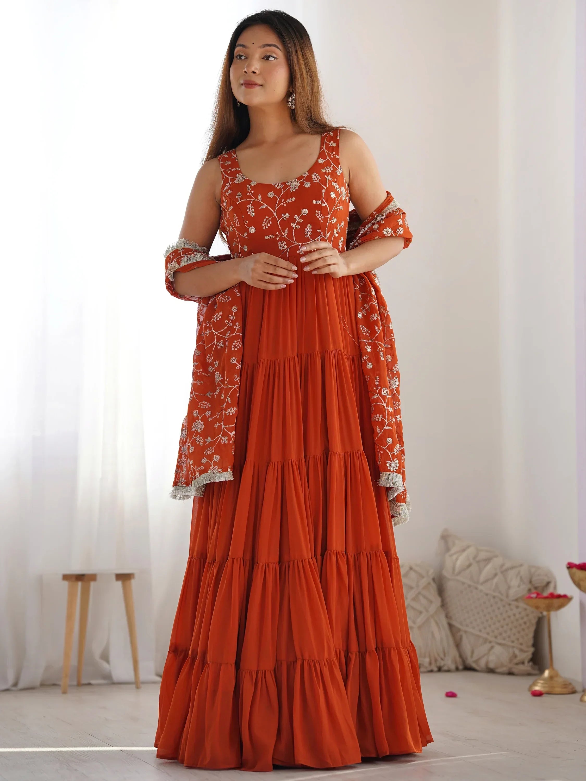 Woman in orange embroidered ethnic gown with matching dupatta, standing indoors