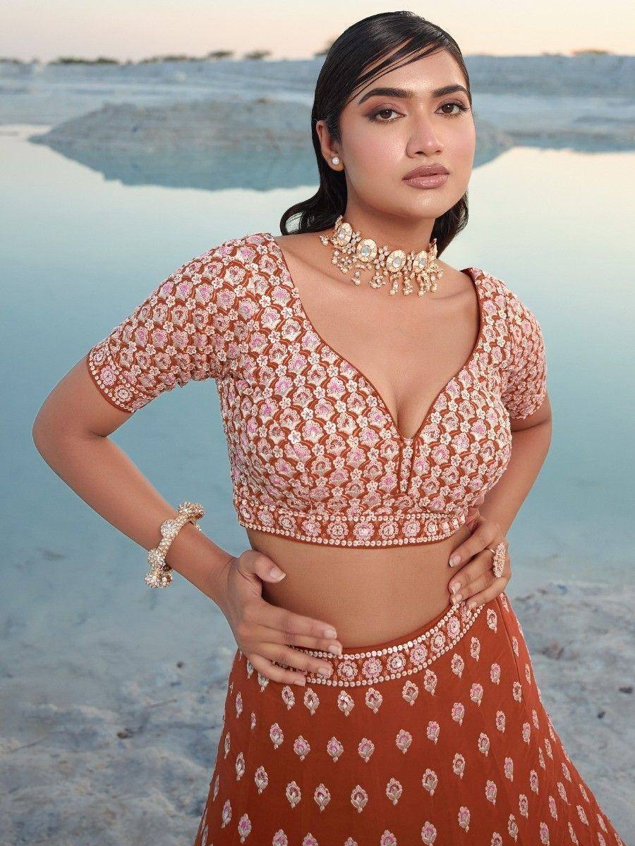 Woman in embroidered terracotta lehenga choli with floral motifs, styled with statement jewelry against a serene water backdrop