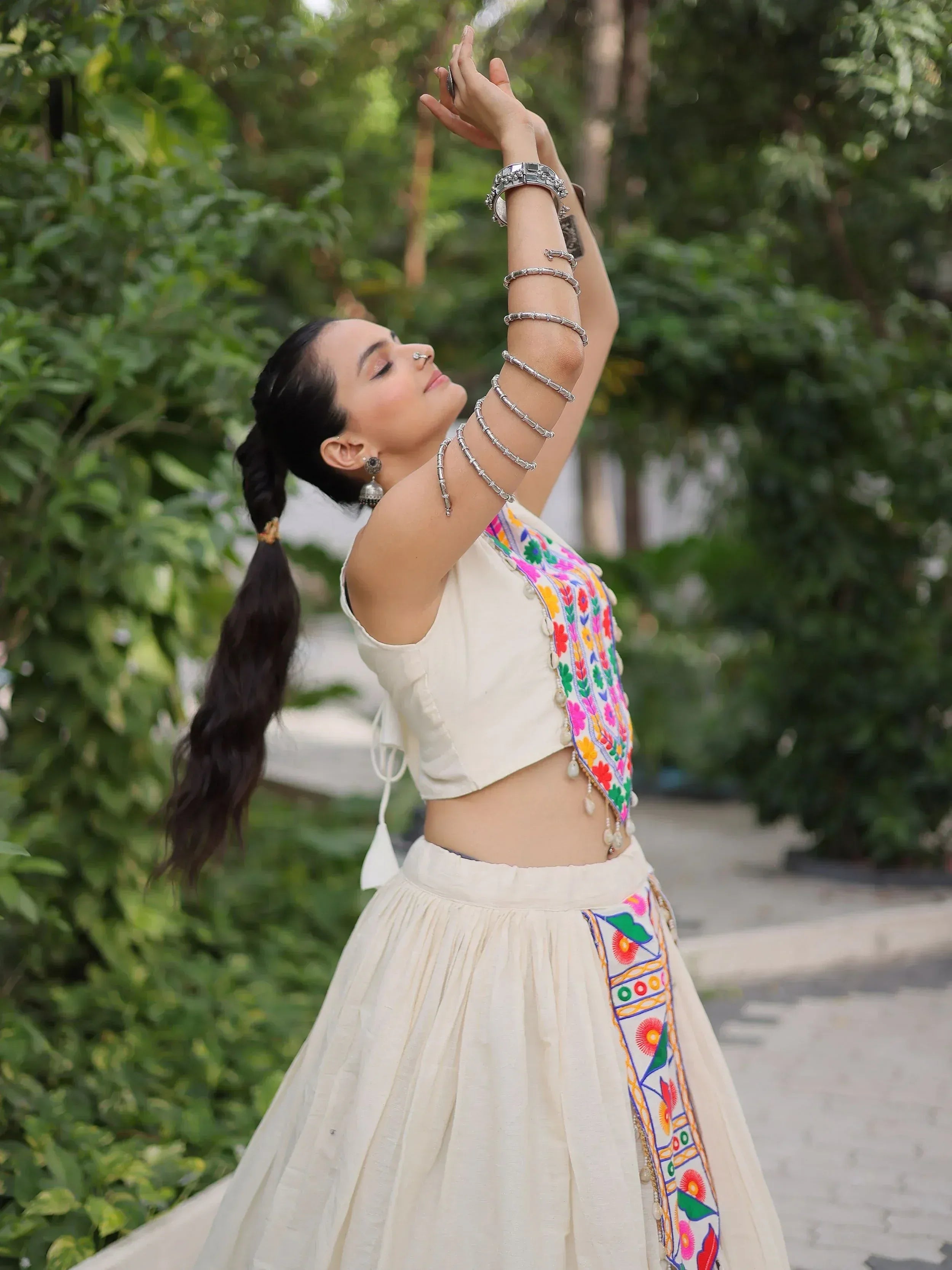 Woman in white embroidered lehenga choli with colorful floral patterns, silver jewelry, outdoors