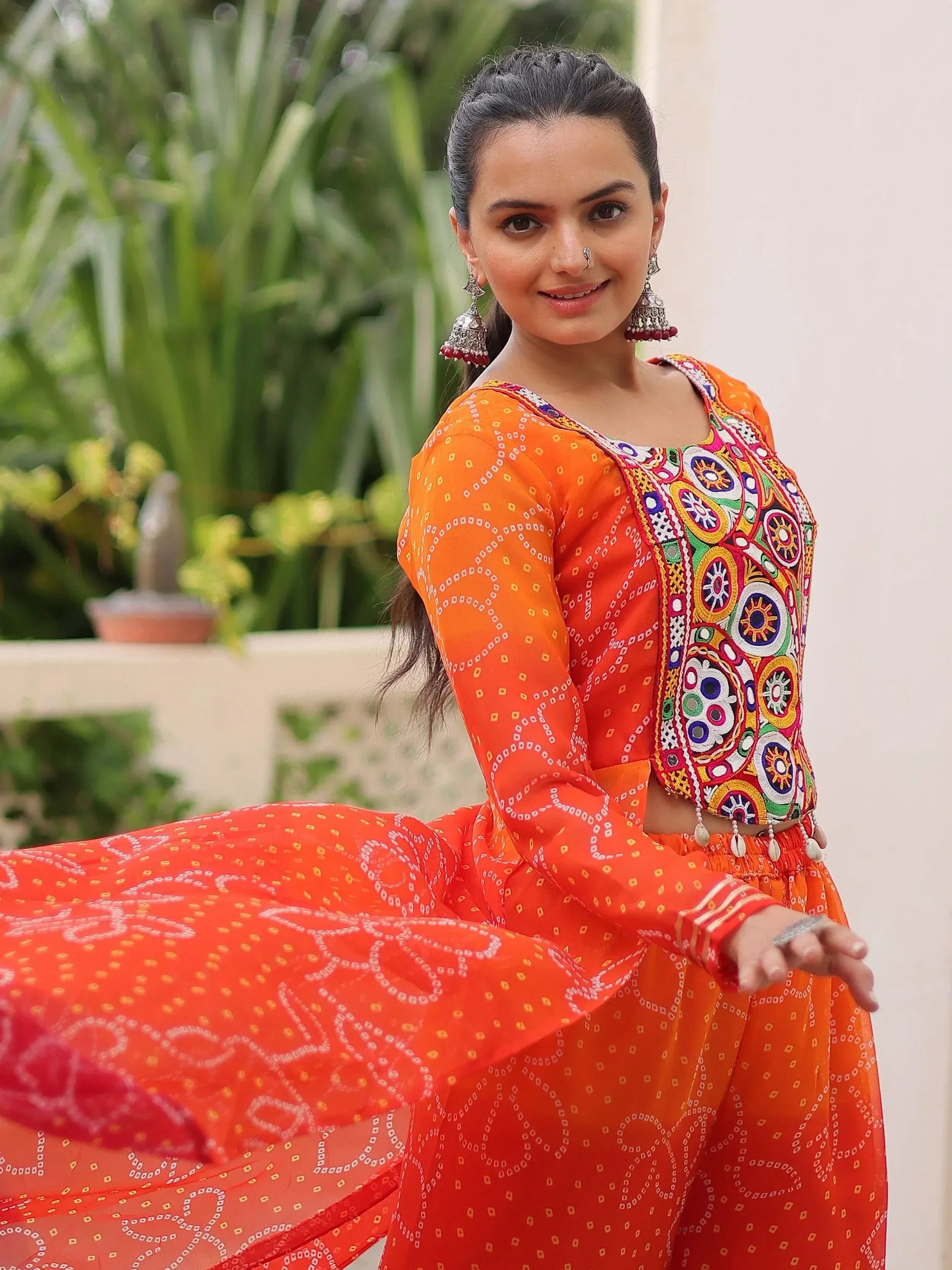 Woman in vibrant orange ethnic wear with colorful mirror work and bandhani print, perfect for festive occasions.