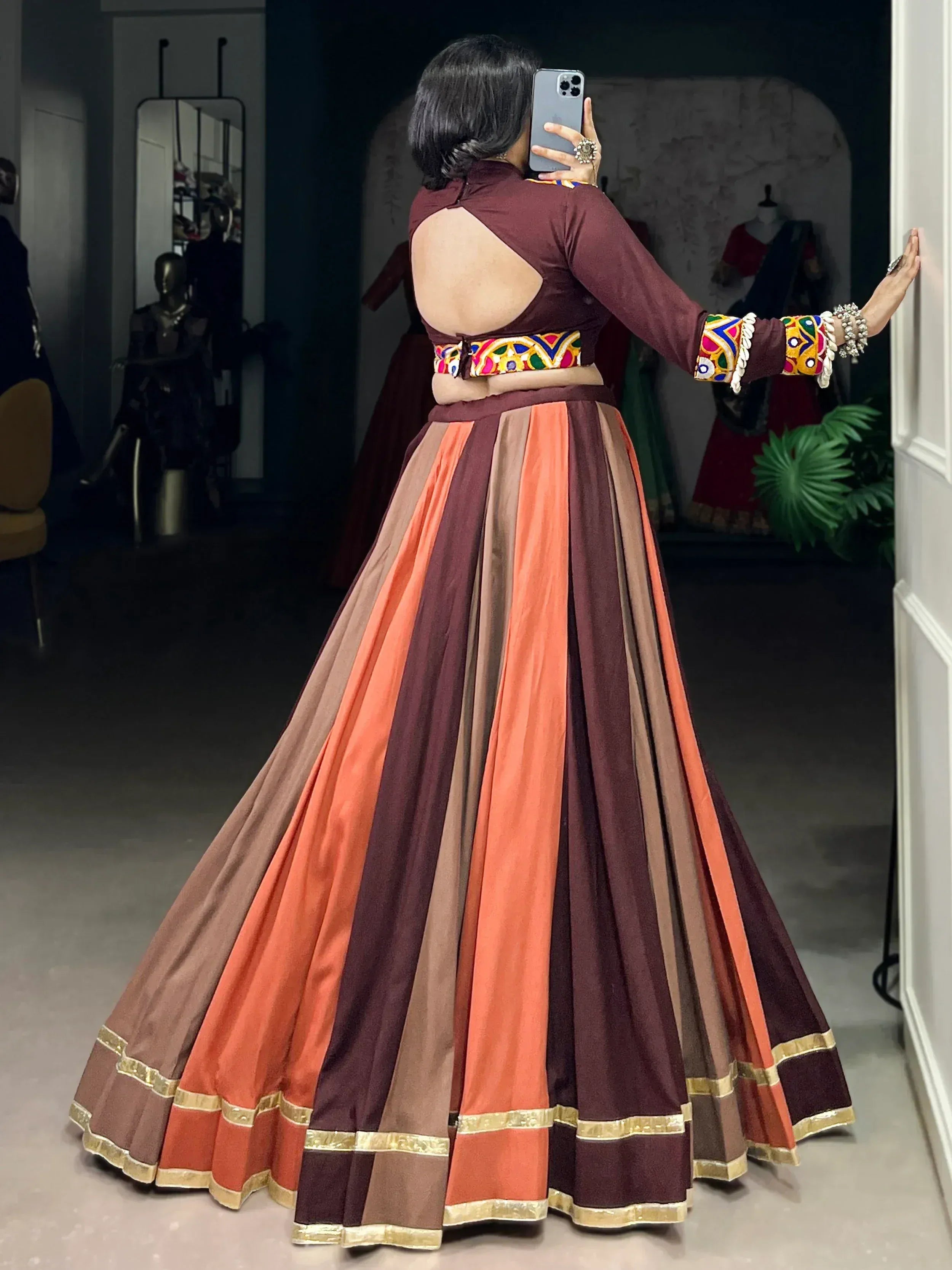 Woman in maroon and orange striped lehenga choli with colorful embroidery, taking mirror selfie in boutique