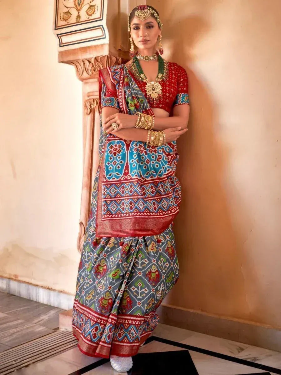 Woman in vibrant Indian saree with red blouse, colorful geometric print, and traditional jewelry, leaning against ornate pillar