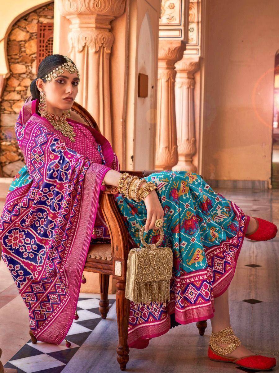 Woman in vibrant Indian ethnic lehenga choli with intricate patterns, gold jewelry, and red flats, seated elegantly.