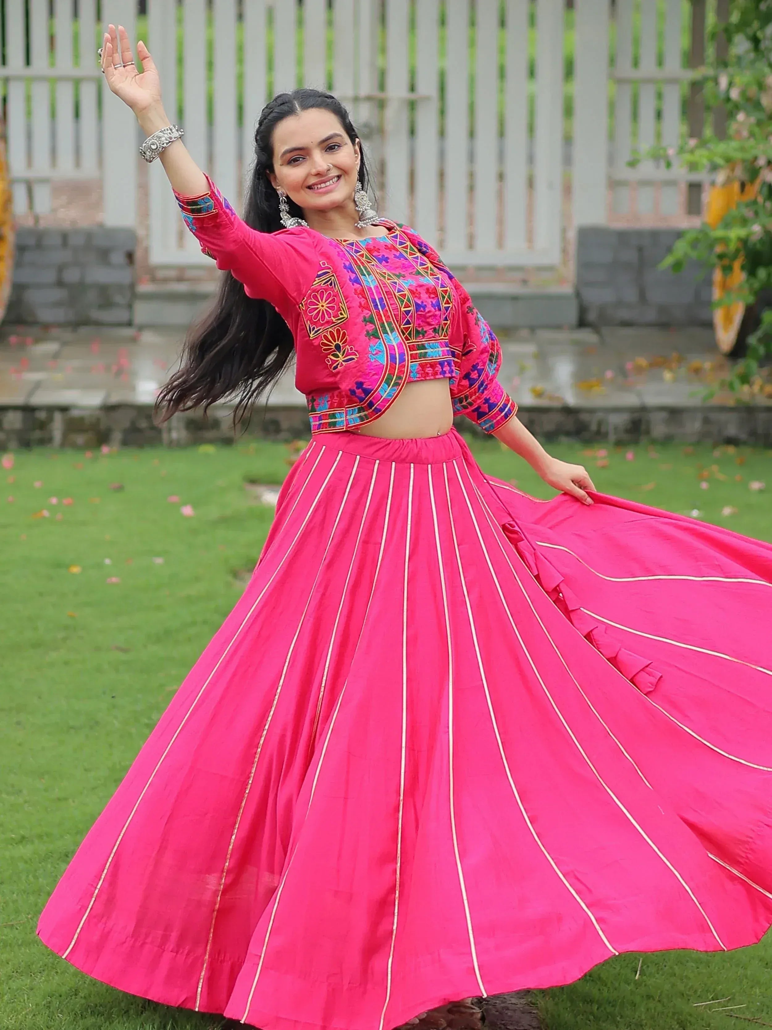 Woman in vibrant pink embroidered lehenga choli twirling outdoors, showcasing ethnic Indian wear with floral patterns and silver jewelry.