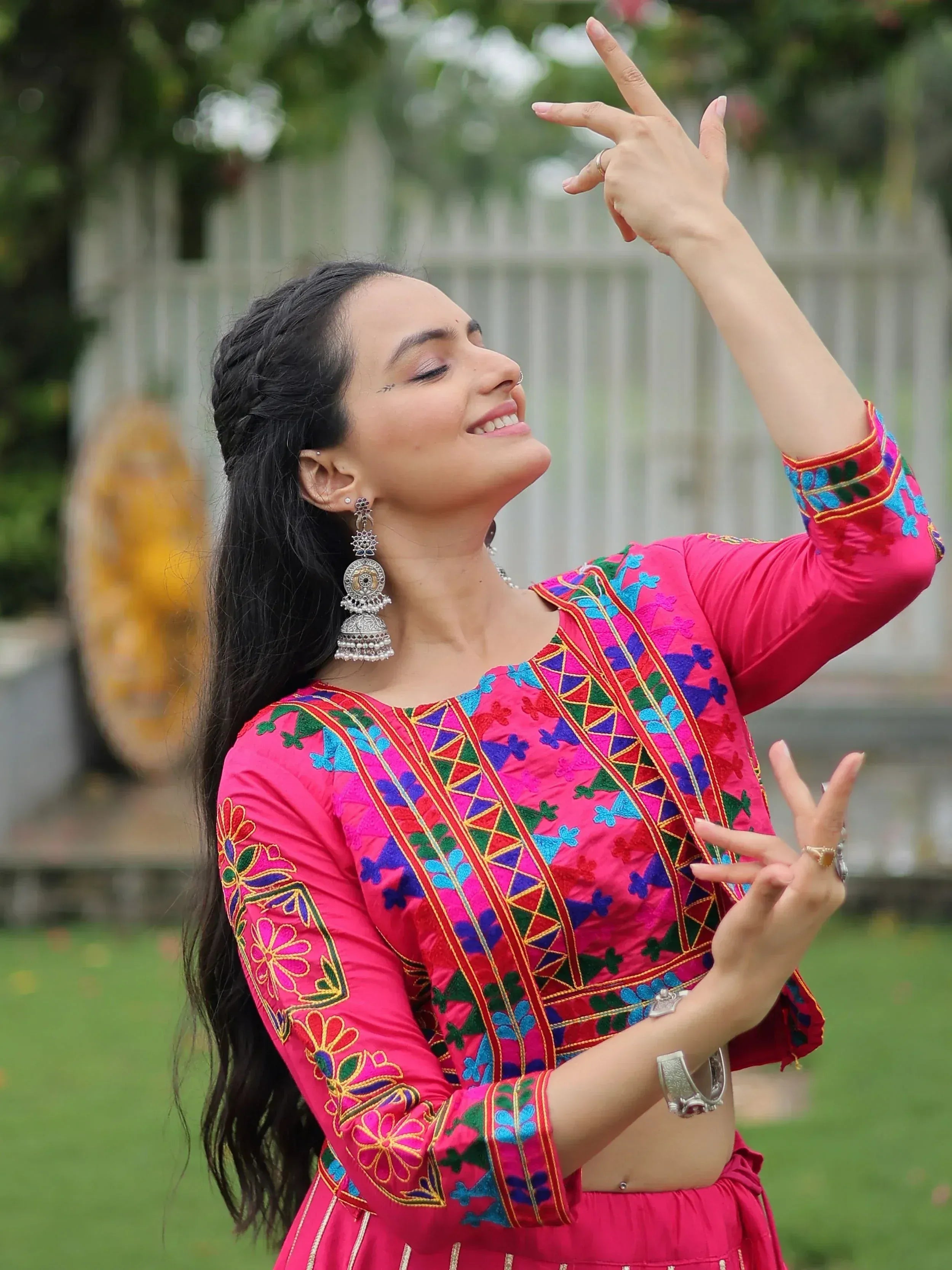 Woman in vibrant pink embroidered ethnic wear, dancing outdoors with traditional jewelry