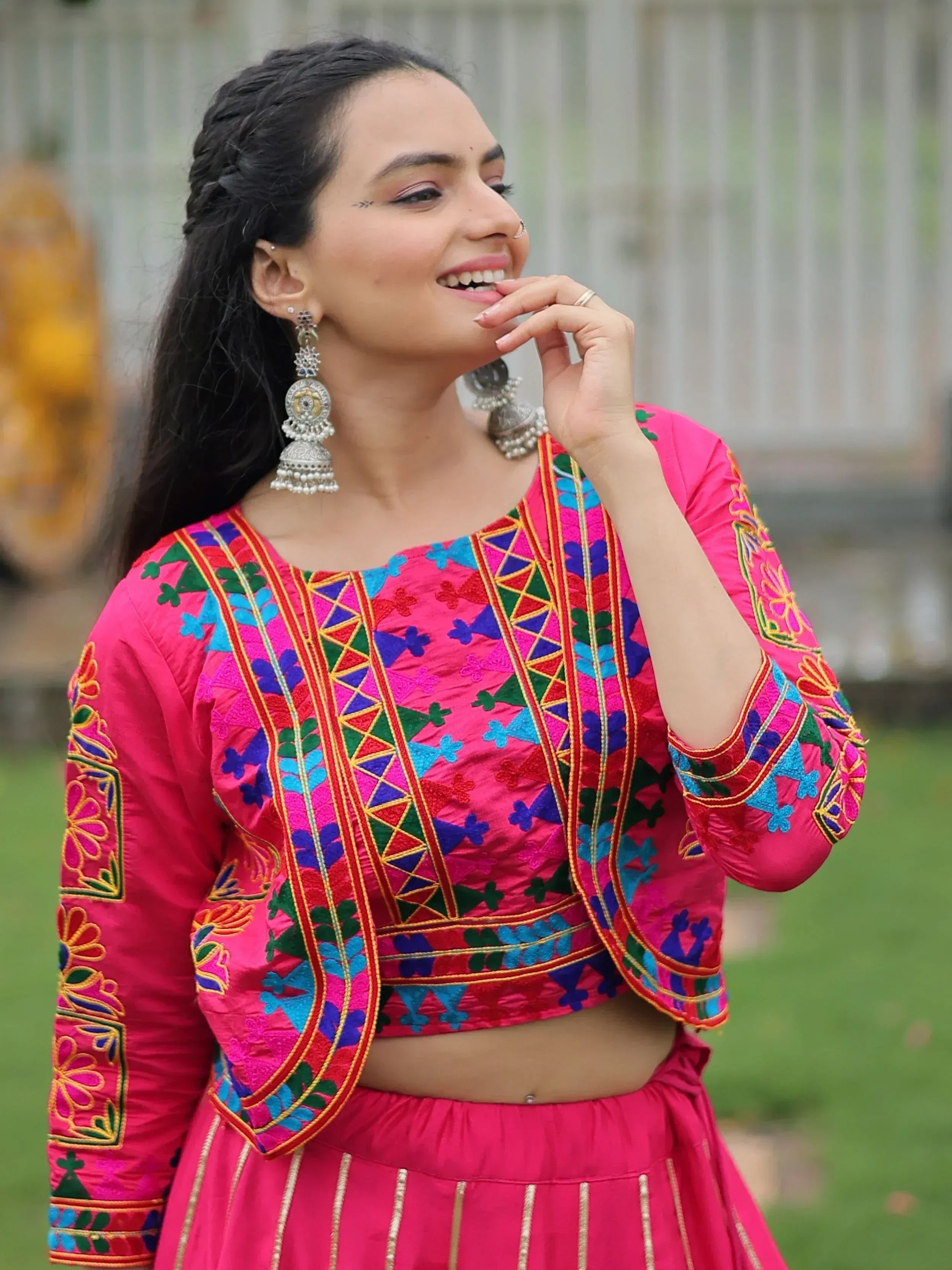 Woman in vibrant pink embroidered lehenga choli with colorful mirror work, styled with jhumka earrings for festive occasions.