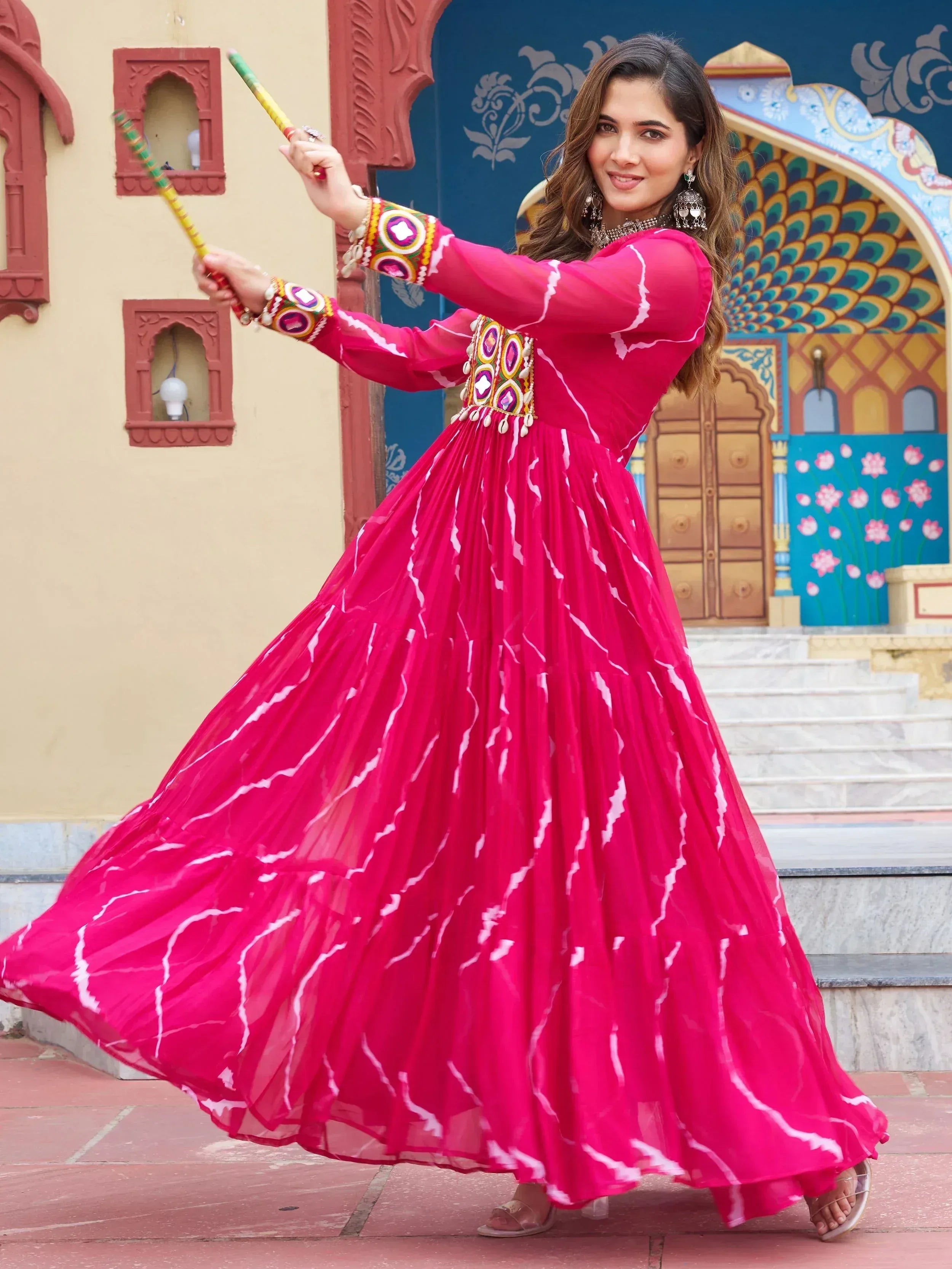 Woman in vibrant pink ethnic gown with mirror work, dancing with dandiya sticks against colorful Indian architecture
