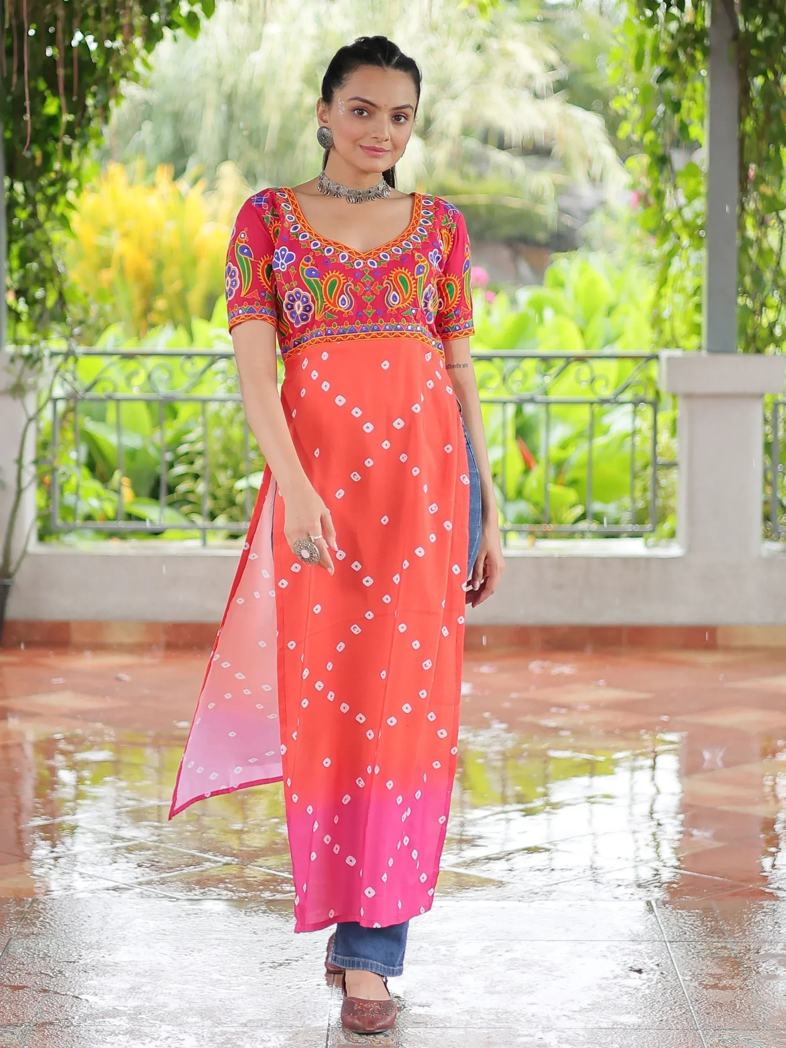 Woman in vibrant red and orange embroidered kurta with bandhani print, paired with jeans, standing on wet patio.
