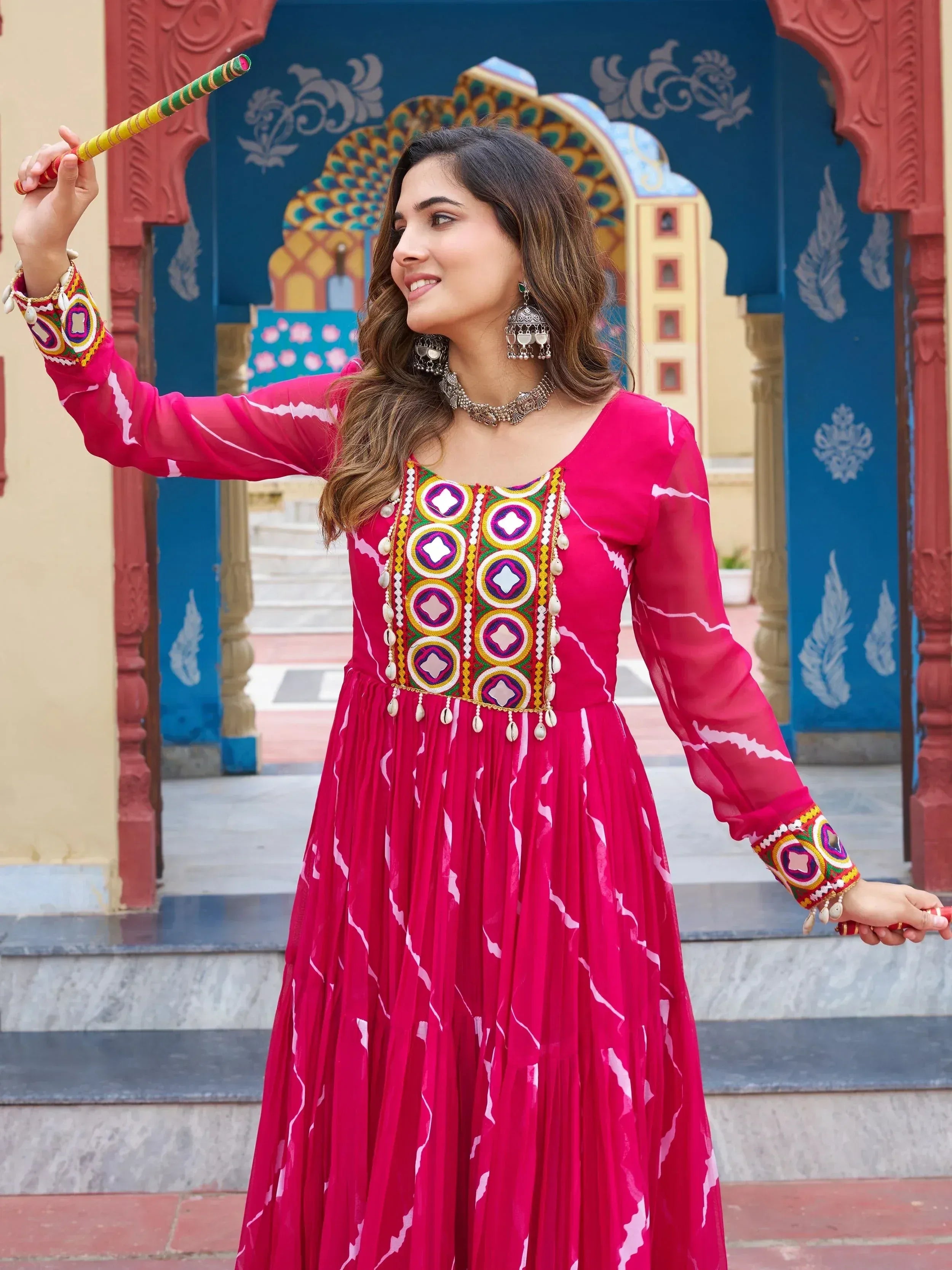 Woman in vibrant pink ethnic gown with mirror work, holding dandiya stick, traditional Indian festive wear
