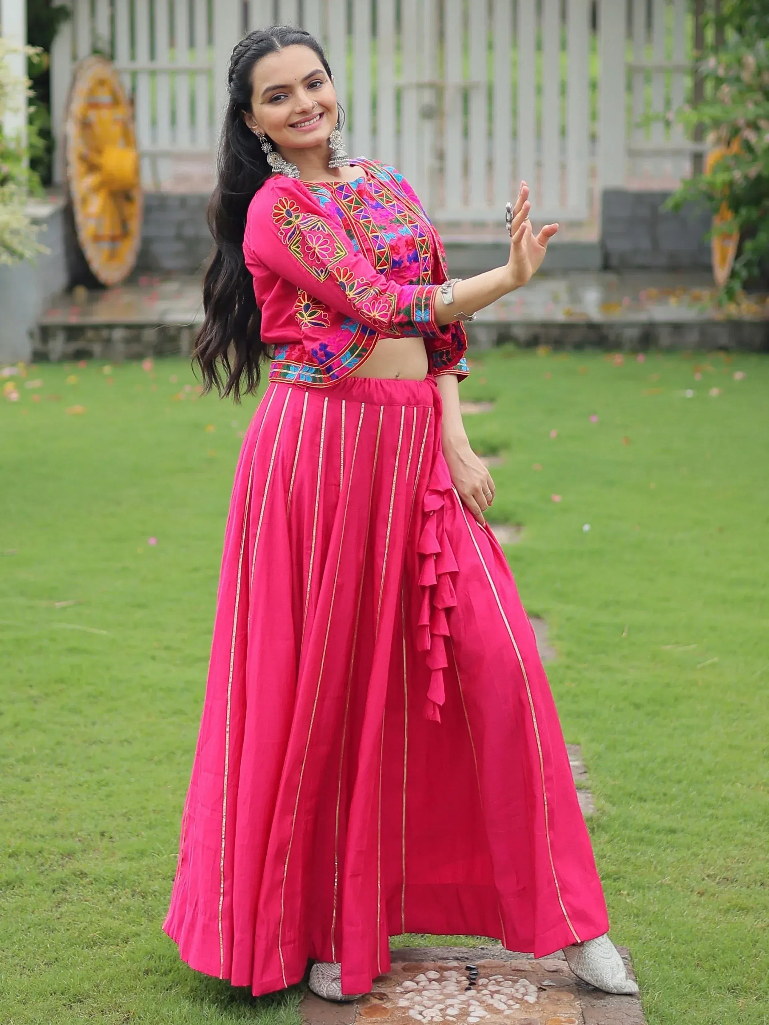 Woman in vibrant pink embroidered lehenga choli with gold stripes, posing outdoors on grass