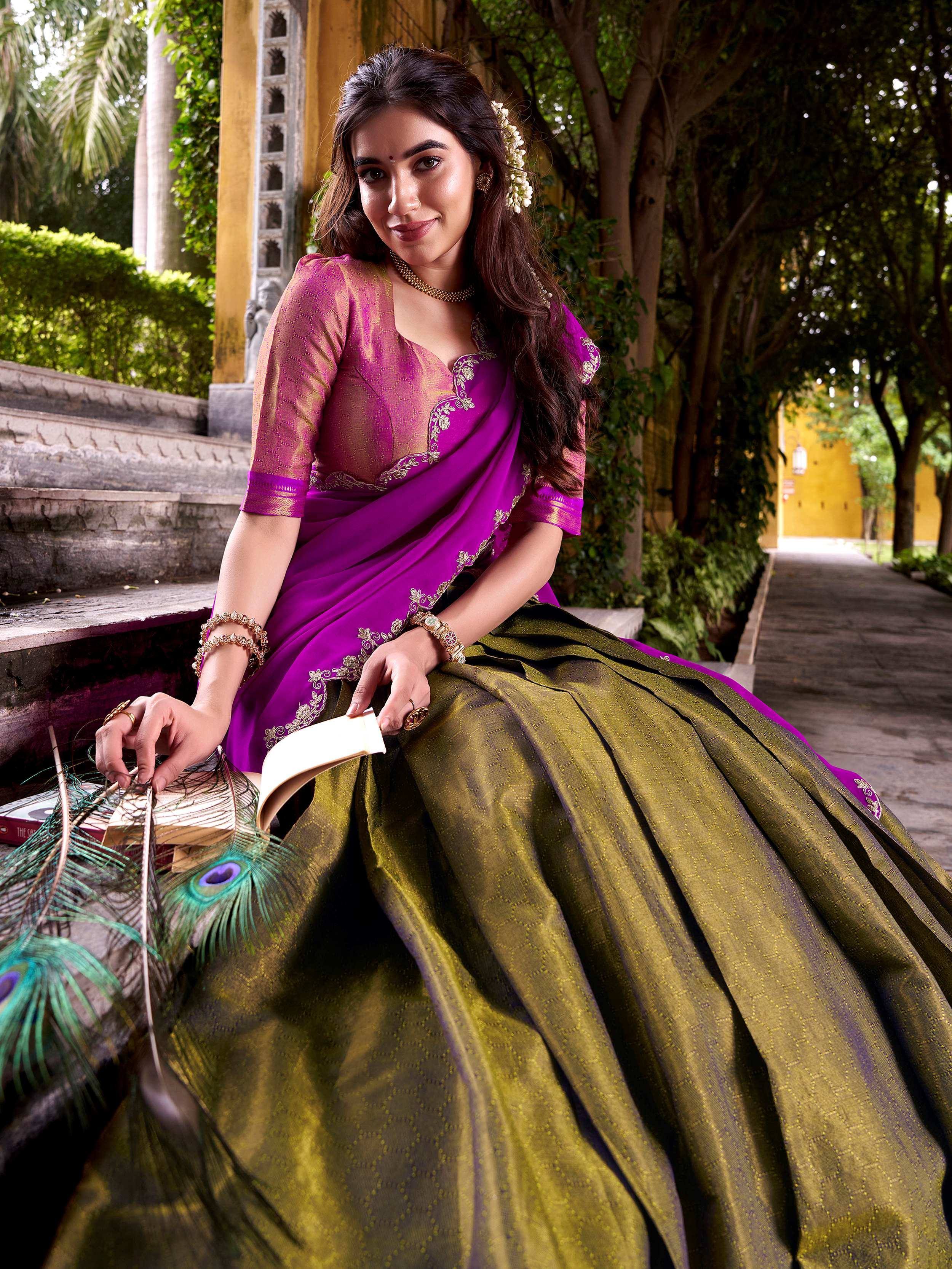 Woman in purple and olive green designer saree with gold embroidery, sitting outdoors with peacock feathers