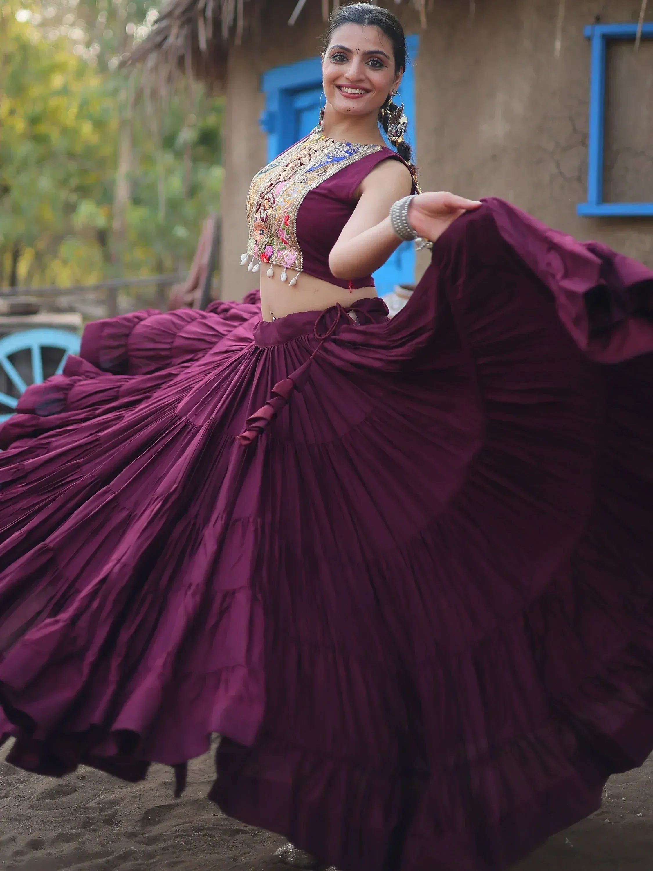 Woman in maroon ethnic lehenga choli with embroidered blouse, twirling outdoors near rustic hut