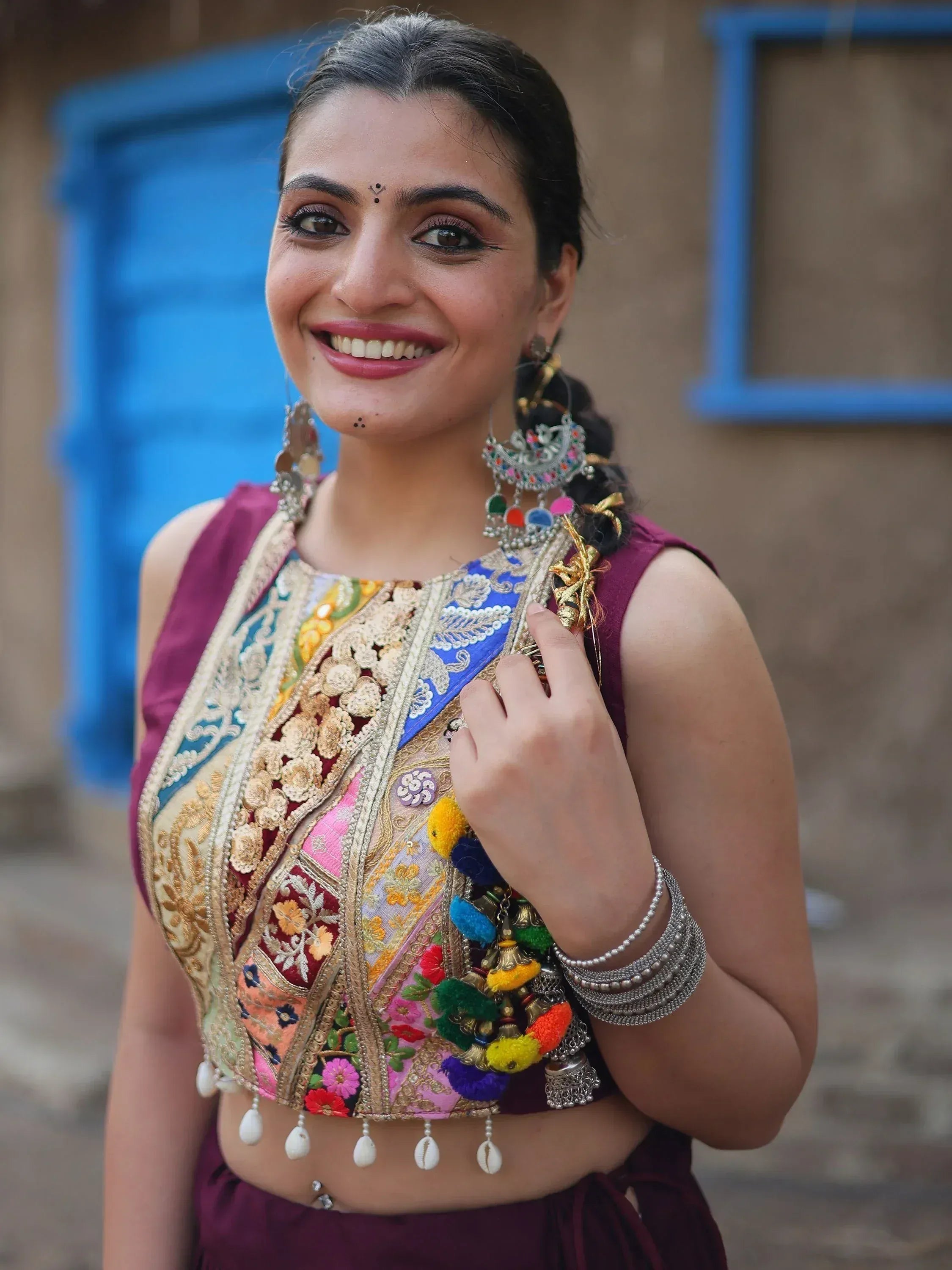 Woman in colorful embroidered ethnic top with pom-poms, shell tassels, and traditional jewelry, smiling outdoors.