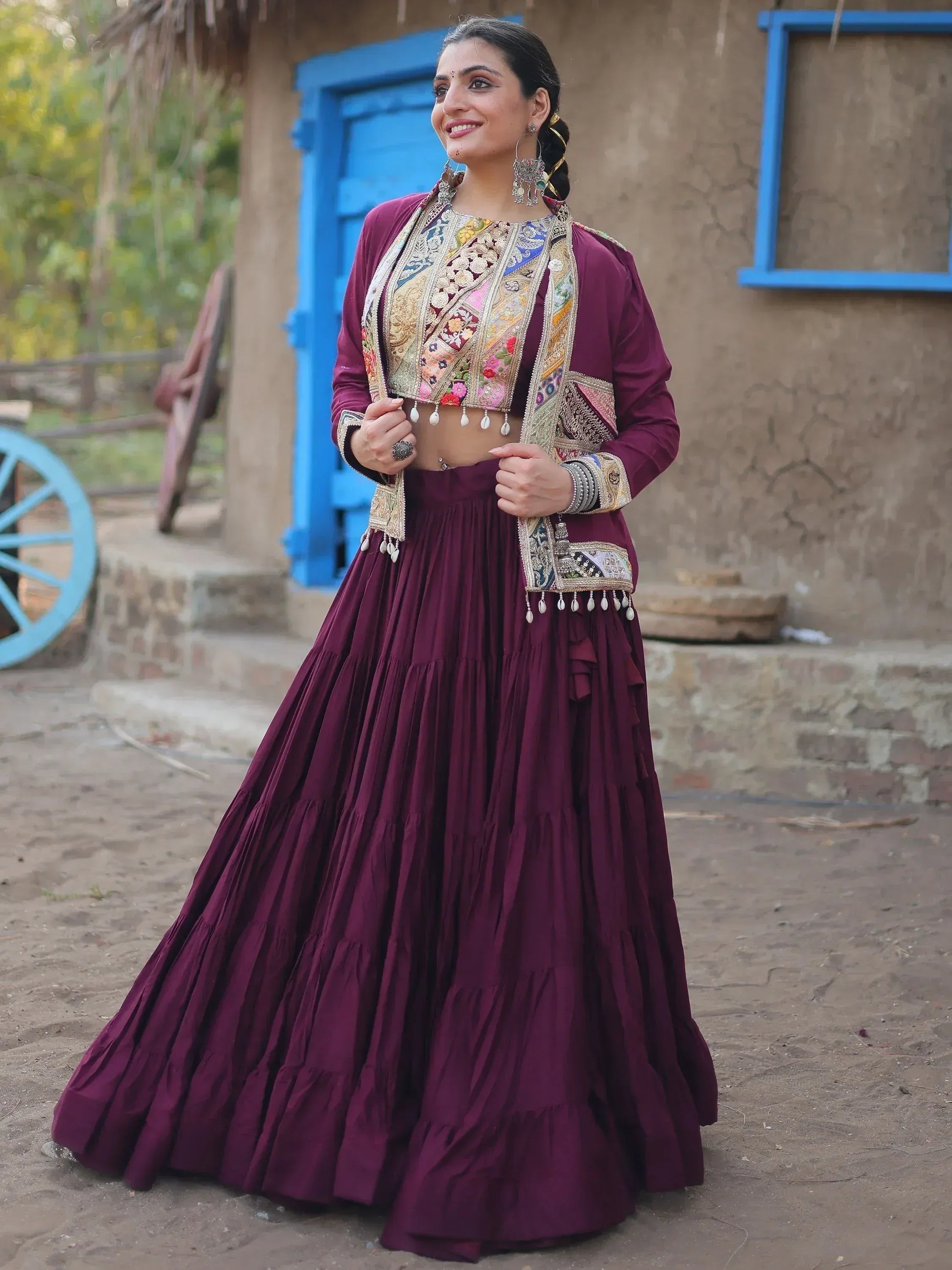 Woman in maroon embroidered lehenga choli with mirror work, standing outdoors near rustic blue door