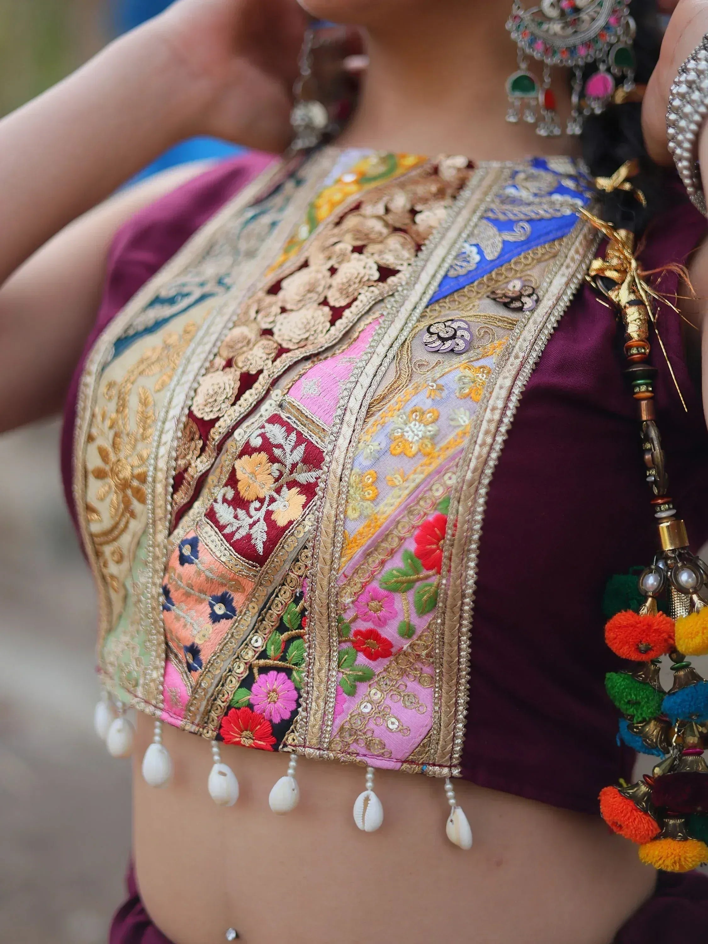 Colorful embroidered ethnic blouse with floral patches, gold trim, and shell tassels, paired with traditional jewelry.