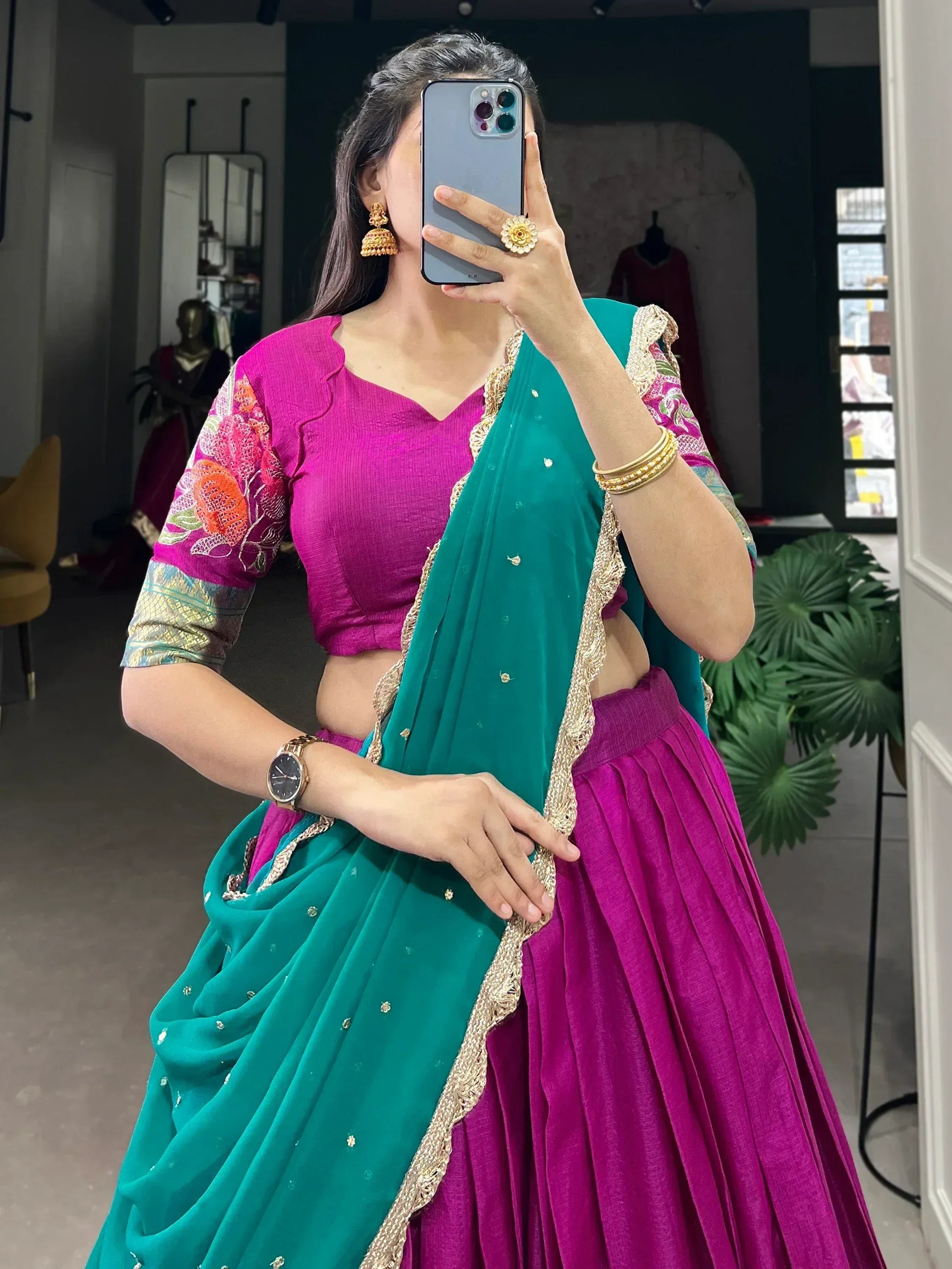 Woman in vibrant pink and green lehenga choli with floral embroidery, gold jewelry, taking mirror selfie