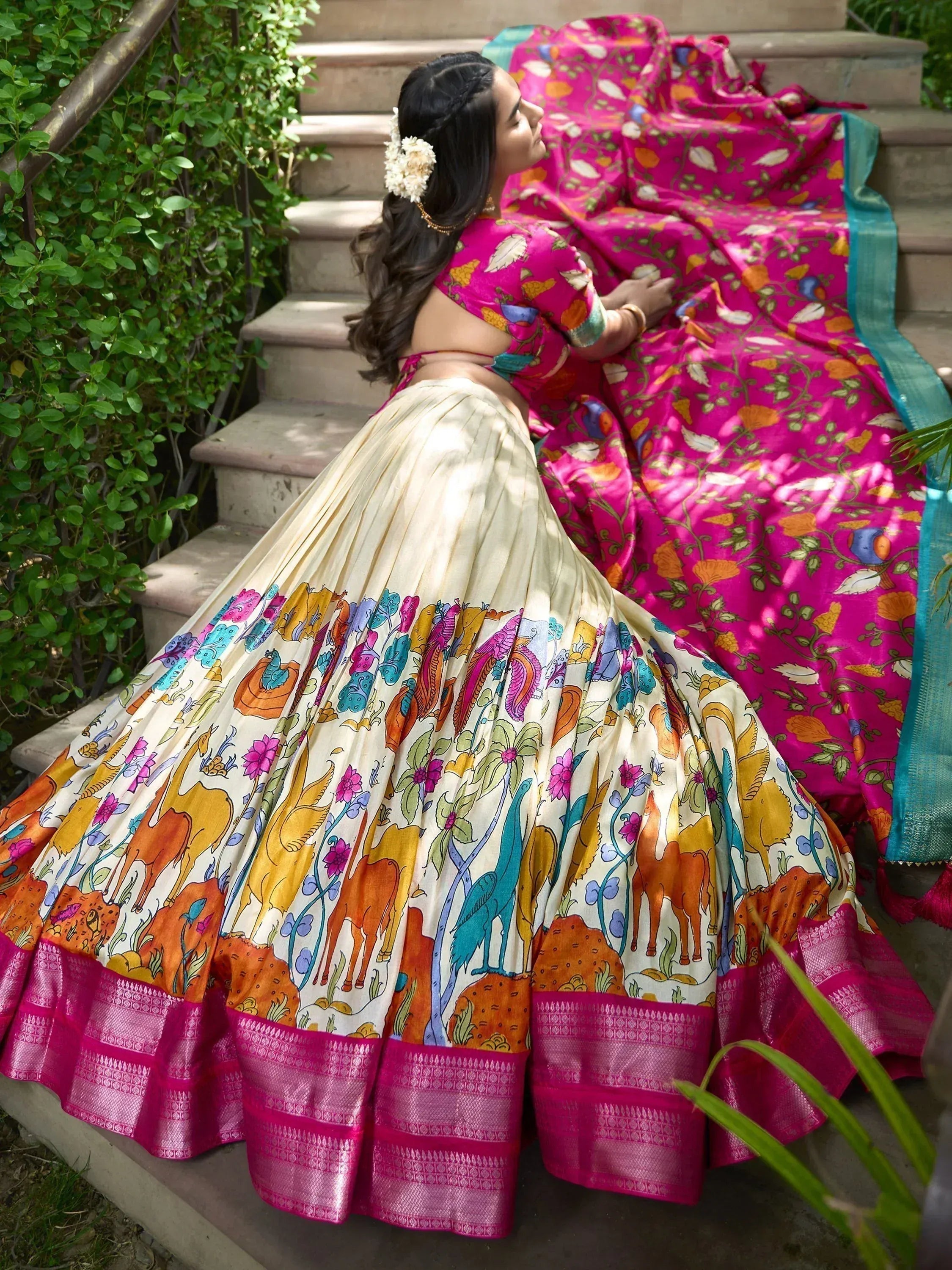 Woman in vibrant pink floral lehenga choli with animal print skirt, sitting on garden steps