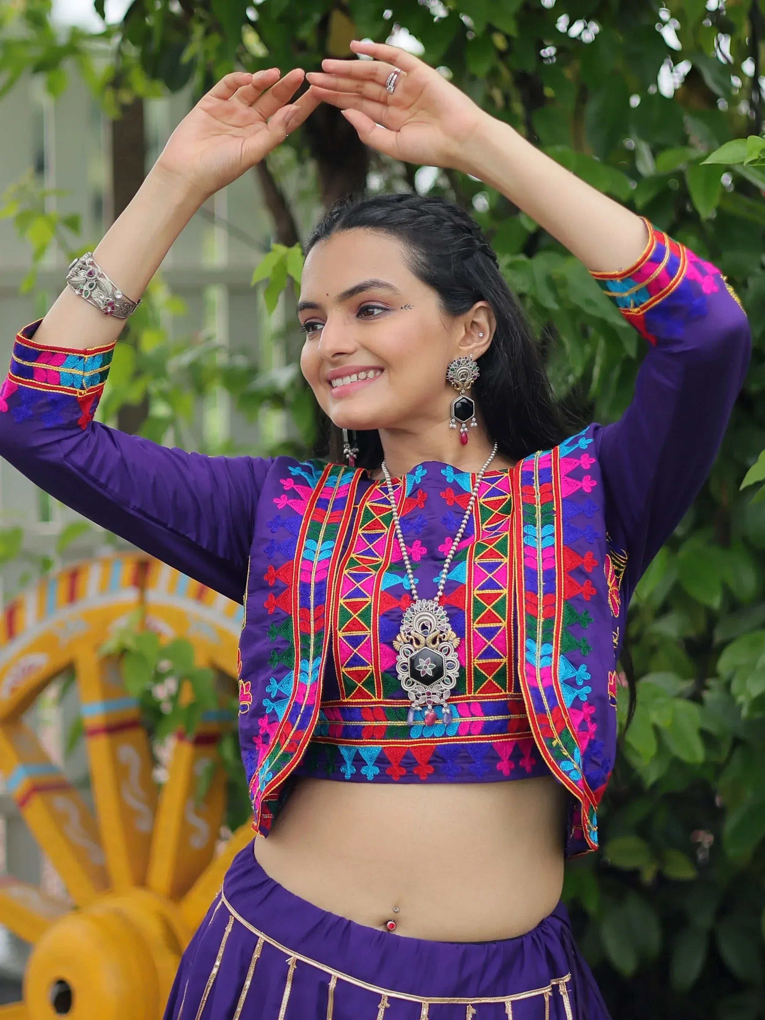 Woman in vibrant purple embroidered ethnic choli with mirror work, paired with matching skirt, traditional jewelry, dancing outdoors.