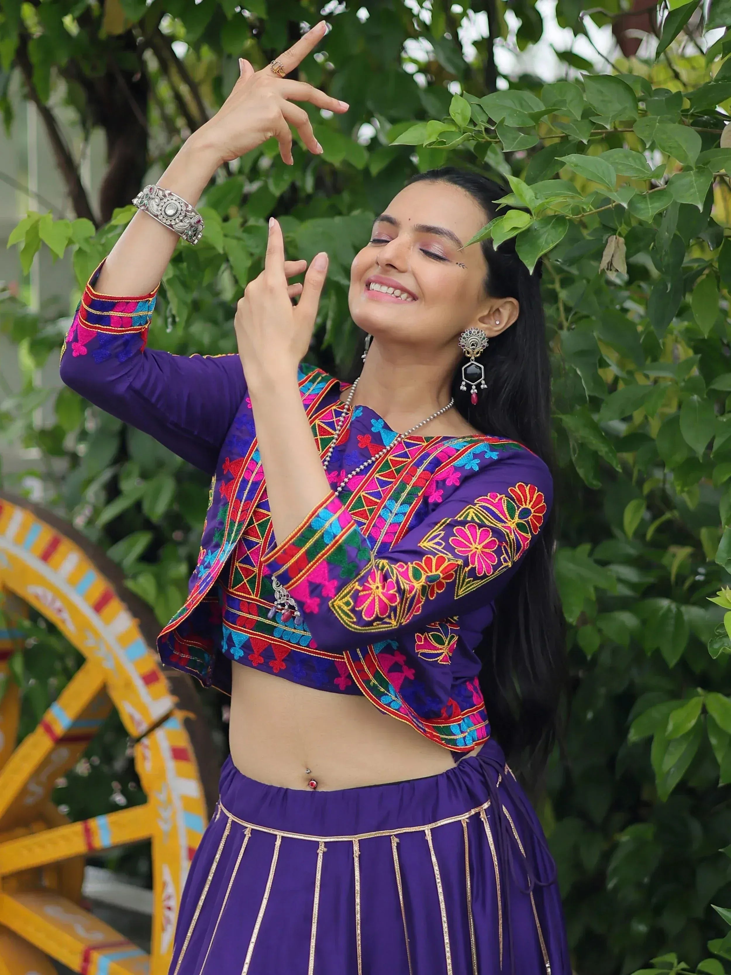 Woman in vibrant purple embroidered ethnic wear with floral patterns, posing outdoors near greenery.