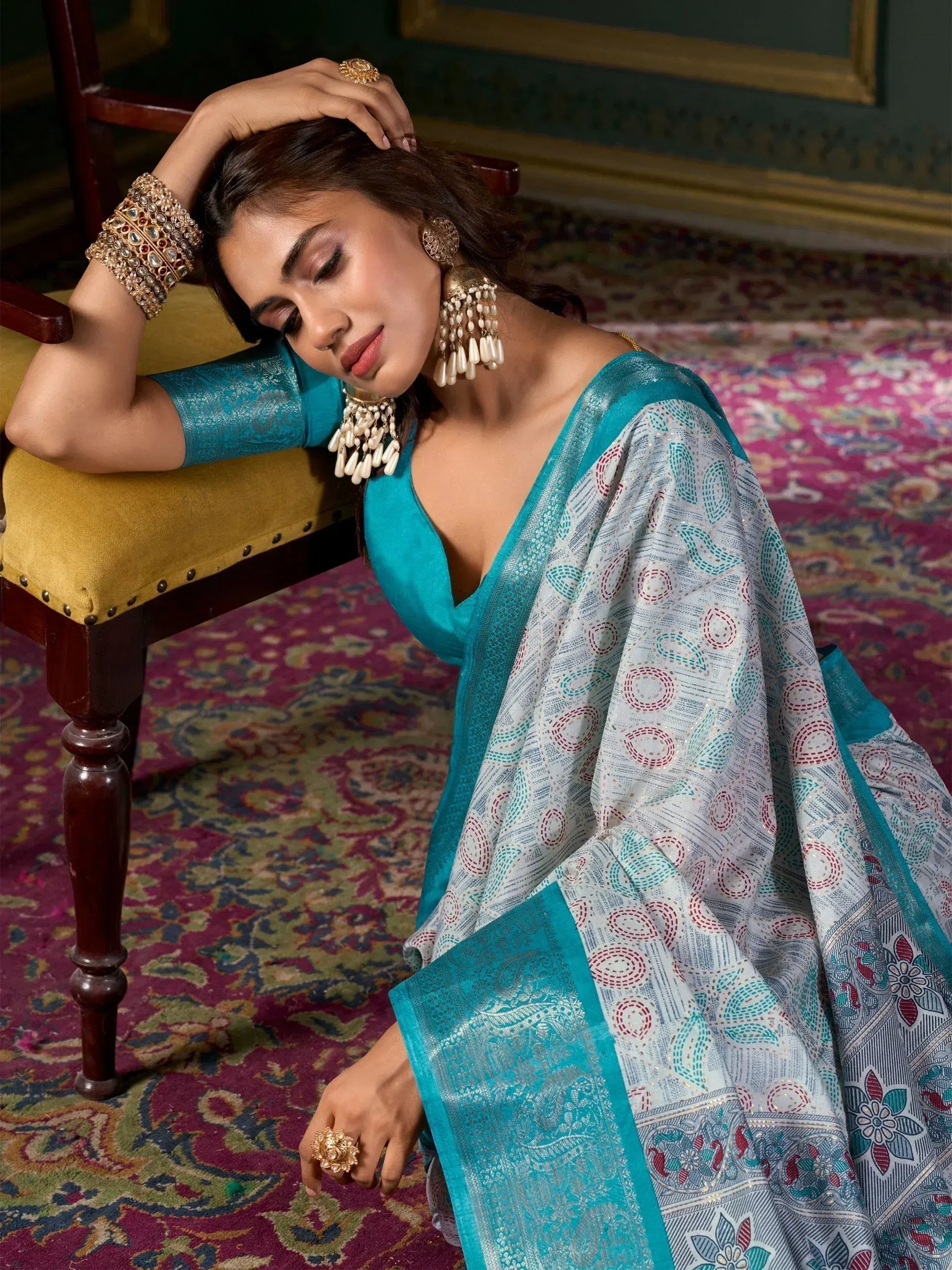 Woman in teal designer saree with printed motifs, traditional jewelry, seated on ornate chair