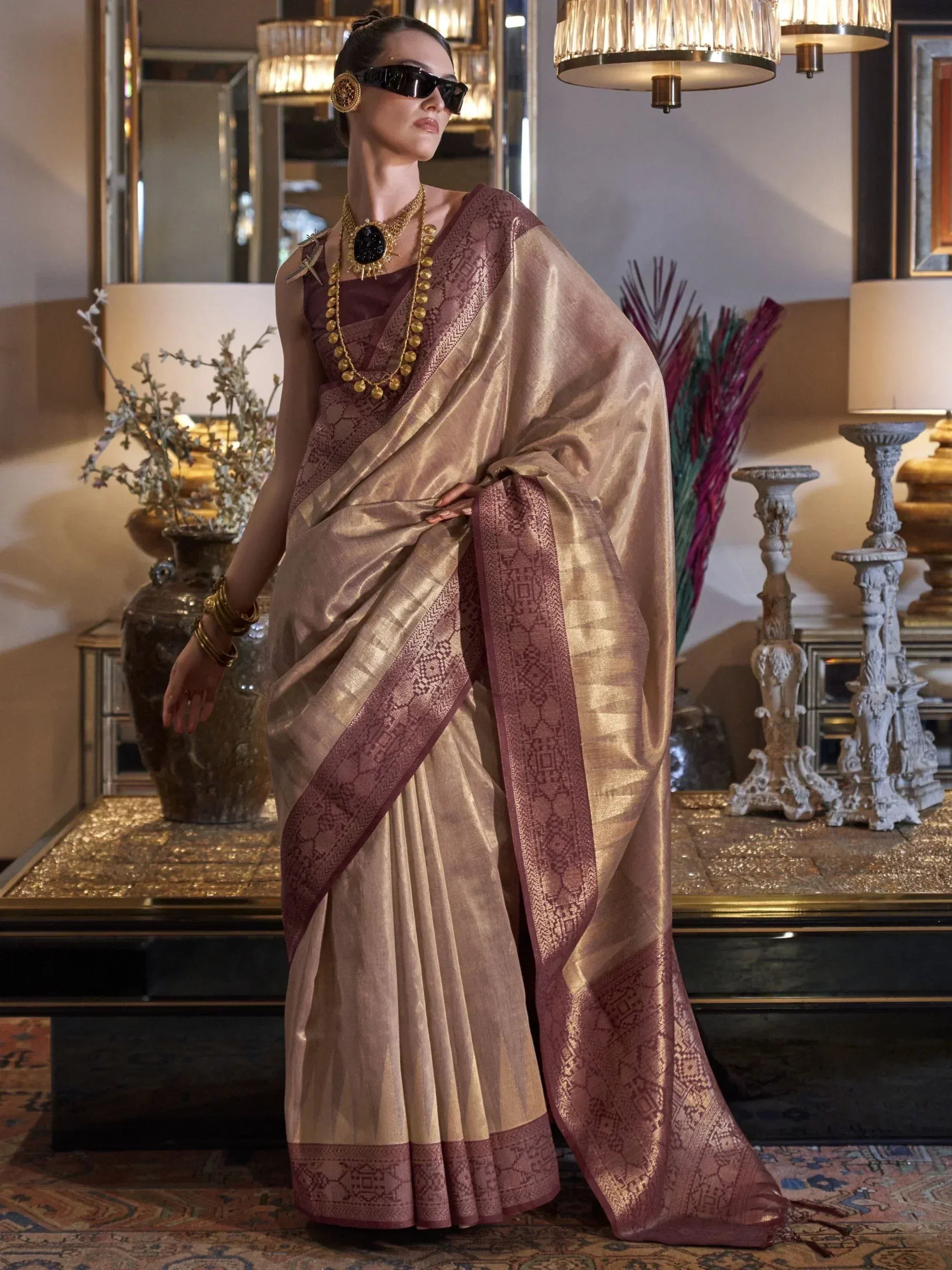 Elegant woman in gold and maroon designer saree with intricate border, styled with bold jewelry and sunglasses.