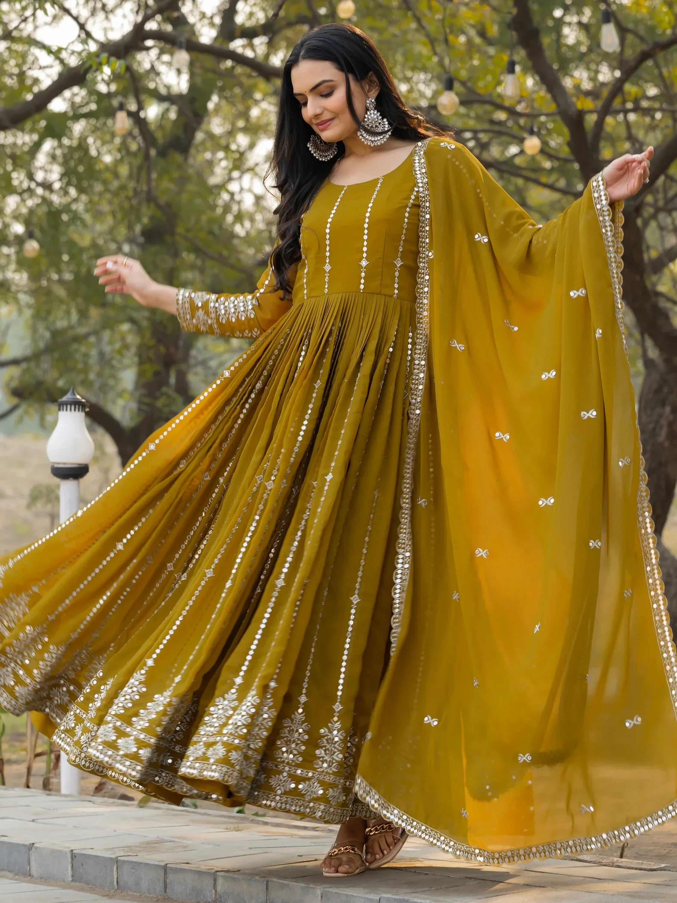 Woman in a mustard yellow embroidered Anarkali gown with dupatta, traditional jewelry, outdoor setting