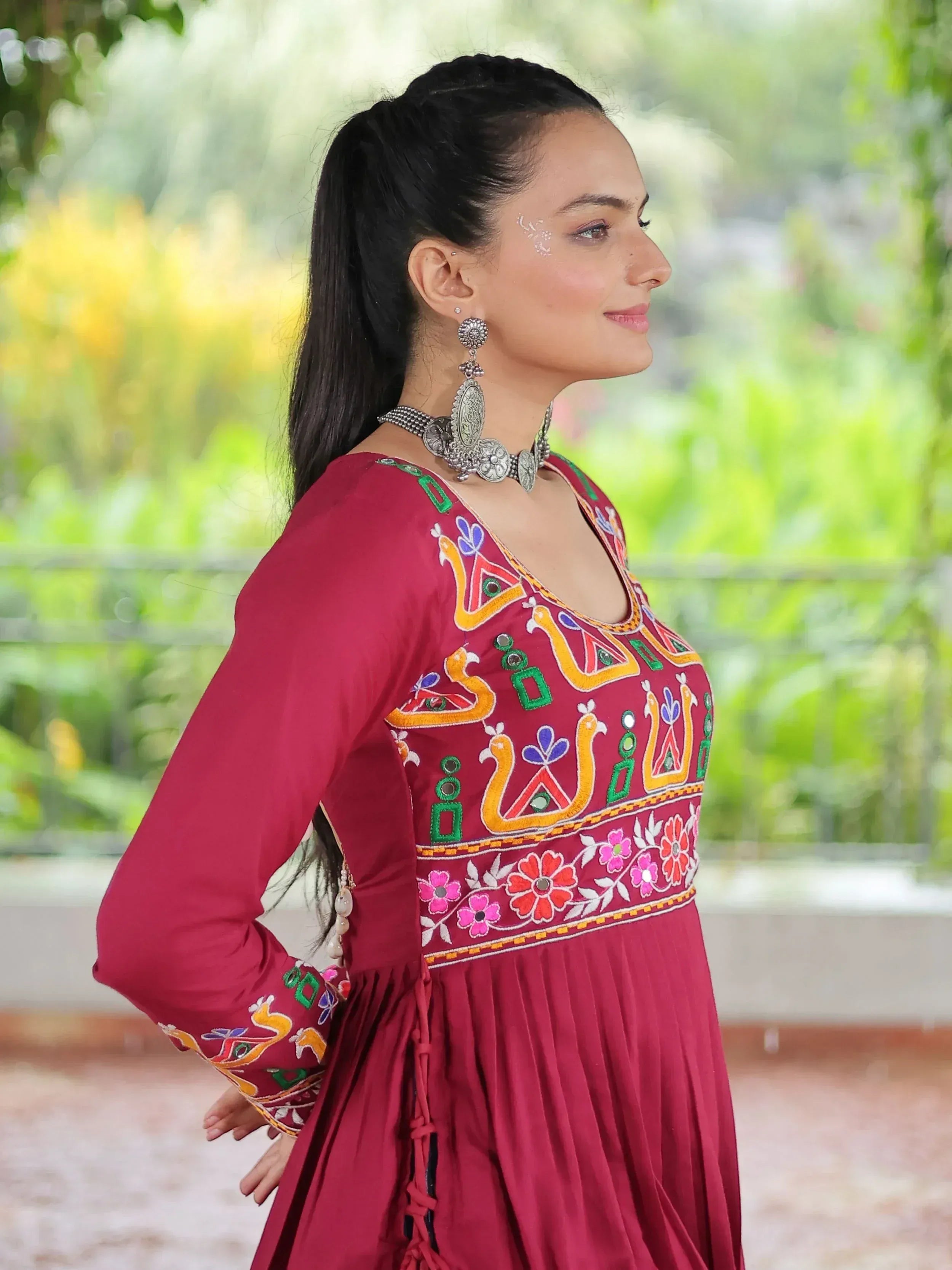 Woman in maroon ethnic gown with colorful embroidery, silver jewelry, and floral patterns, perfect for Indian festive wear.