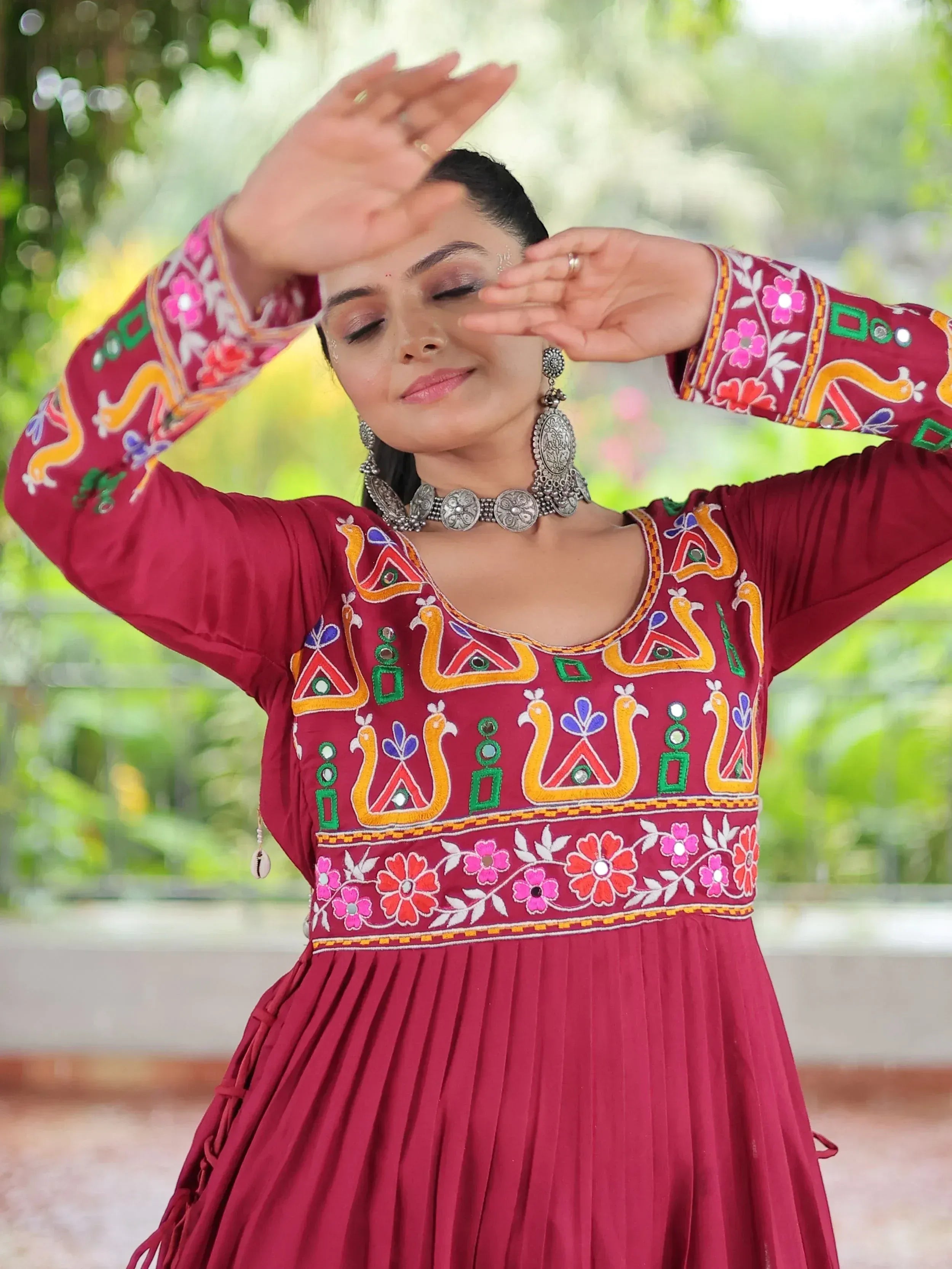 Woman in maroon ethnic gown with colorful embroidery, floral patterns, and mirror work, styled with traditional jewelry.