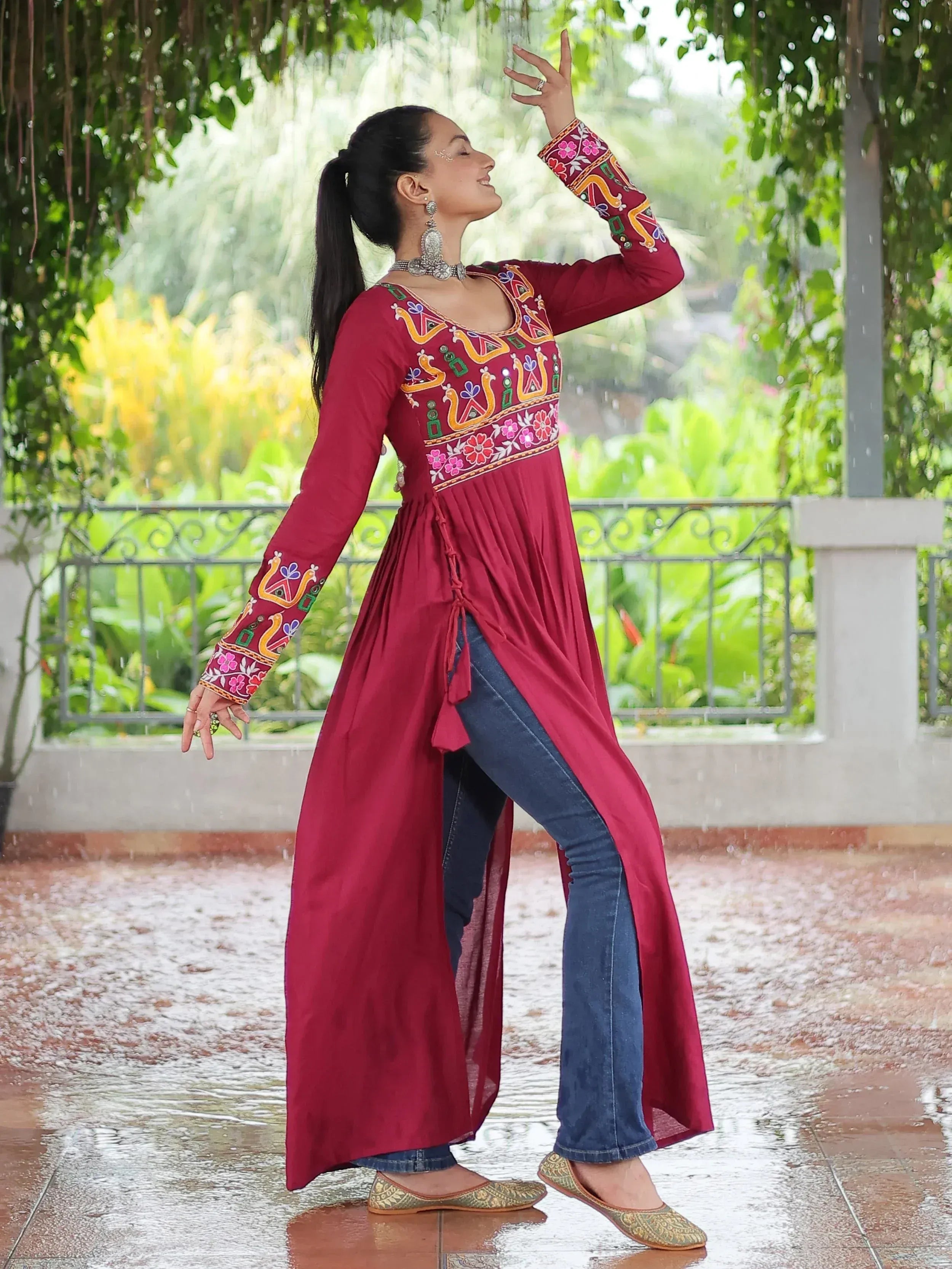 Woman in maroon embroidered ethnic kurti with jeans, enjoying rain under greenery