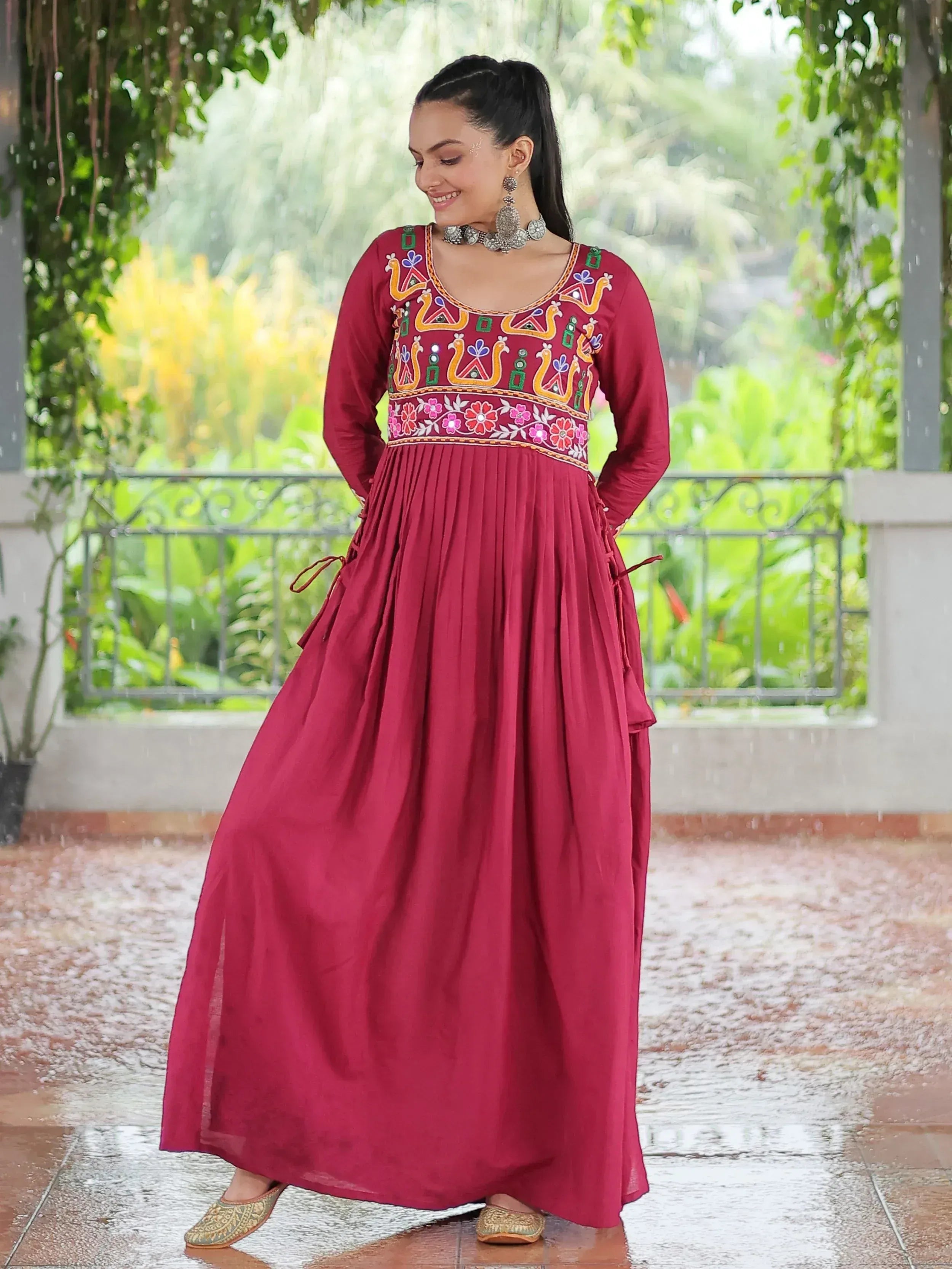 Maroon ethnic gown with colorful embroidery, floral motifs, and pleated skirt, paired with traditional jewelry and juttis.
