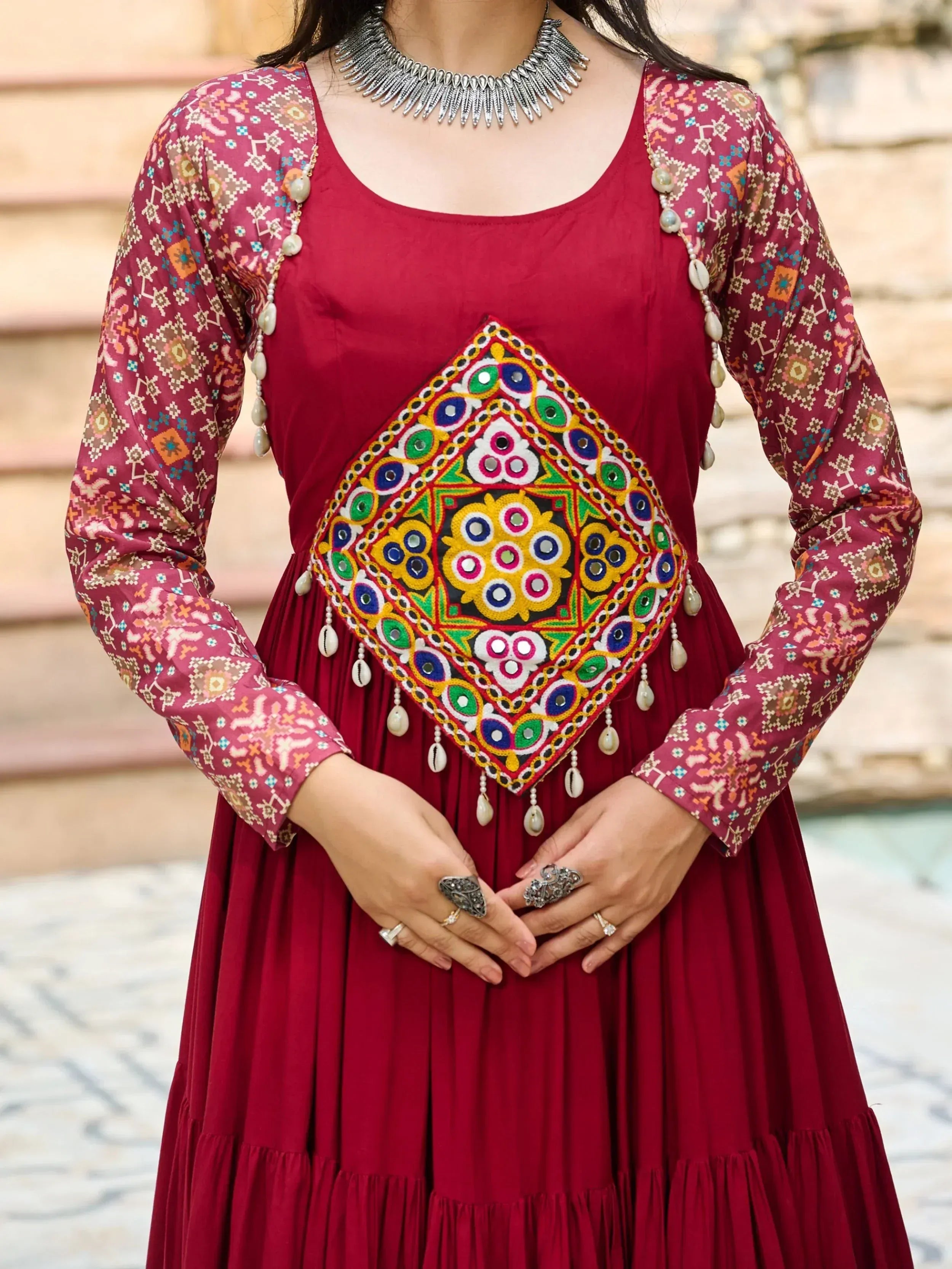 Maroon ethnic gown with mirror work, printed sleeves, and shell tassels, paired with statement jewelry