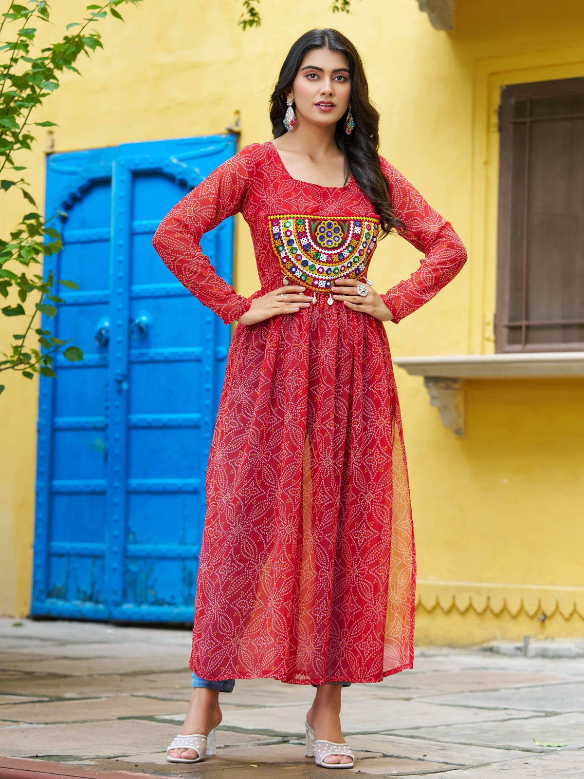Red ethnic long dress with colorful mirror work, worn with denim shorts and white heels against yellow wall and blue door