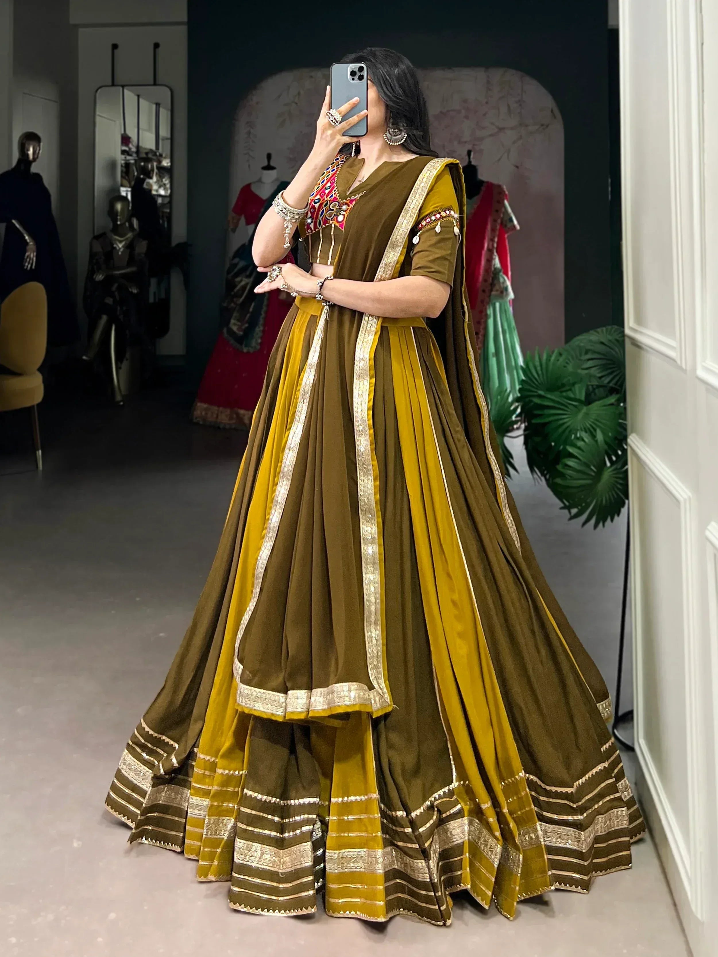 Woman in olive and mustard Indian lehenga choli with gold embroidery, taking mirror selfie in boutique