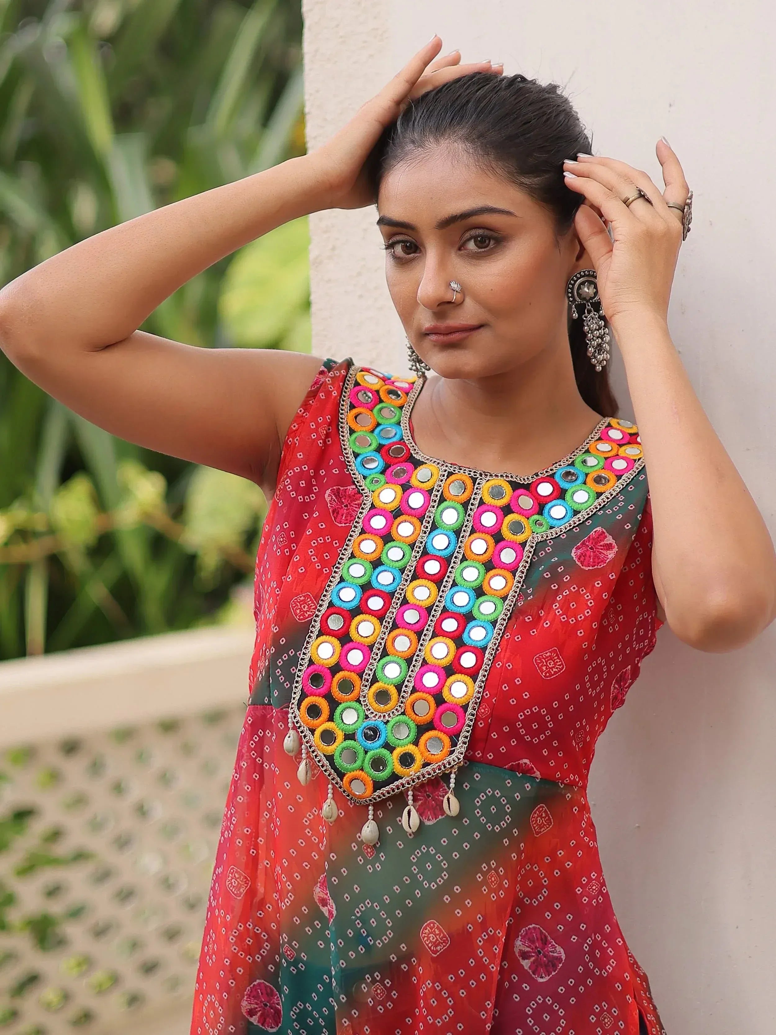 Woman in vibrant red and green ethnic dress with colorful mirror work and floral print, styled with traditional jewelry.