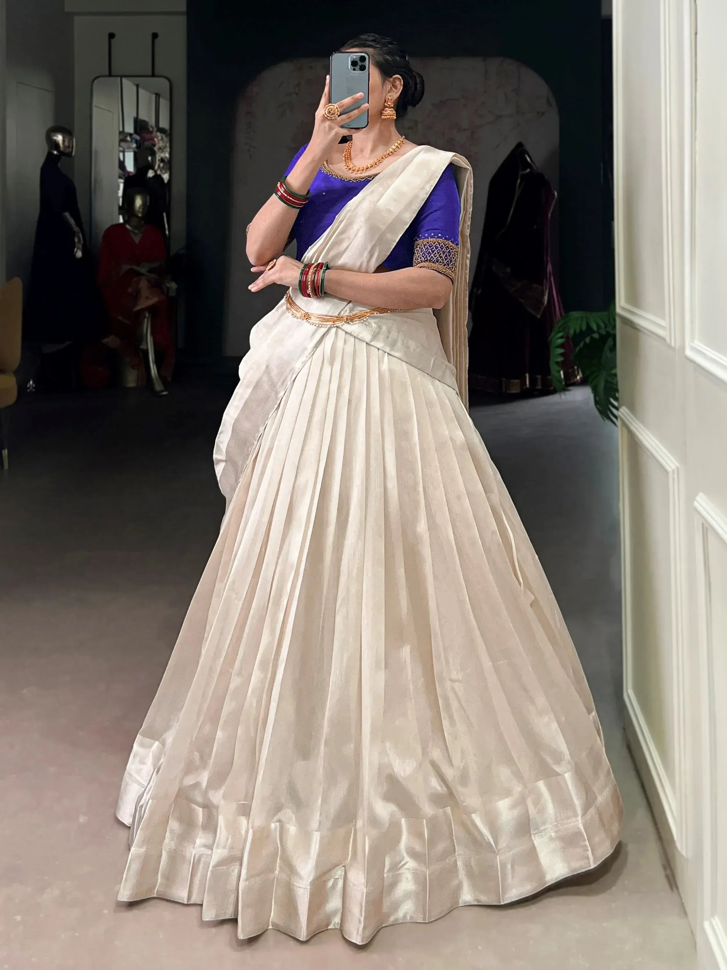 Woman in cream pleated lehenga with blue blouse, gold jewelry, taking mirror selfie in boutique