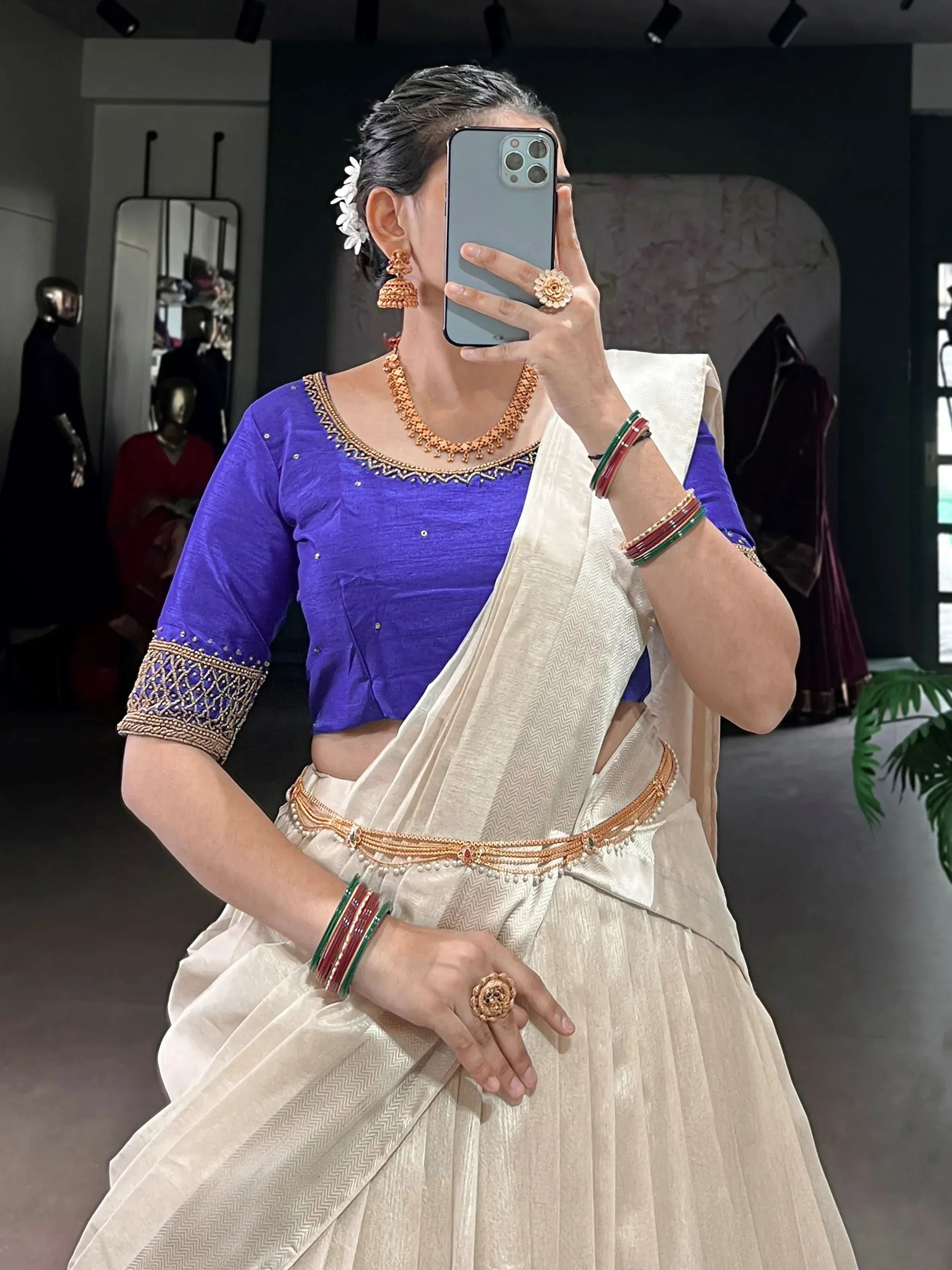 Woman in purple blouse and cream saree with gold jewelry, taking mirror selfie in boutique