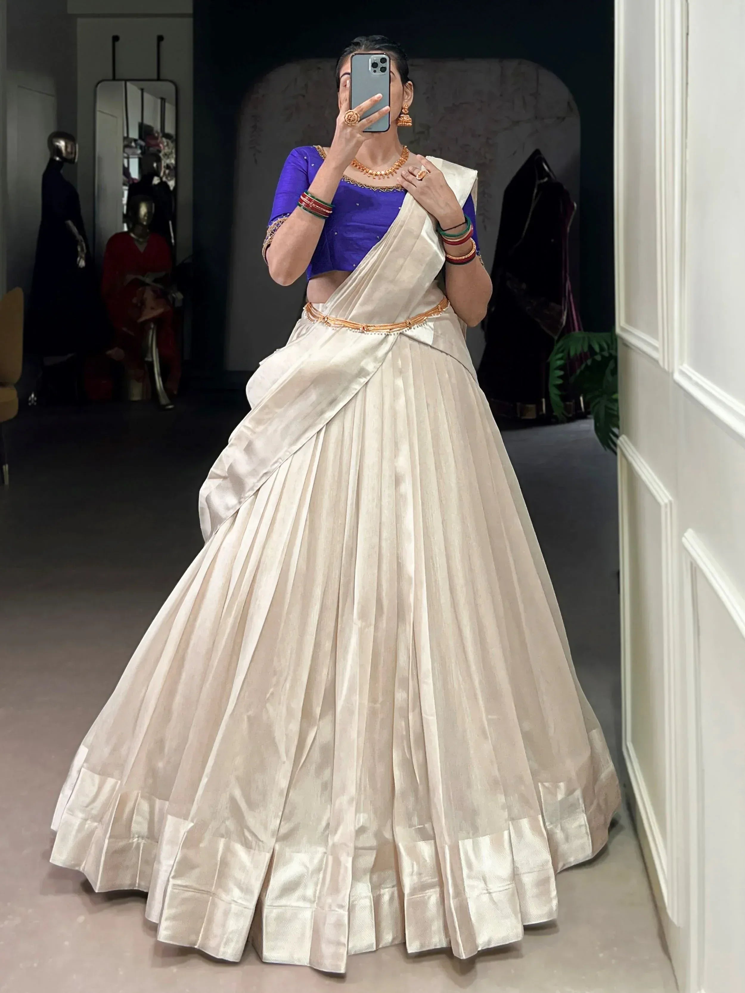 Woman in cream pleated lehenga with blue blouse, gold jewelry, taking mirror selfie