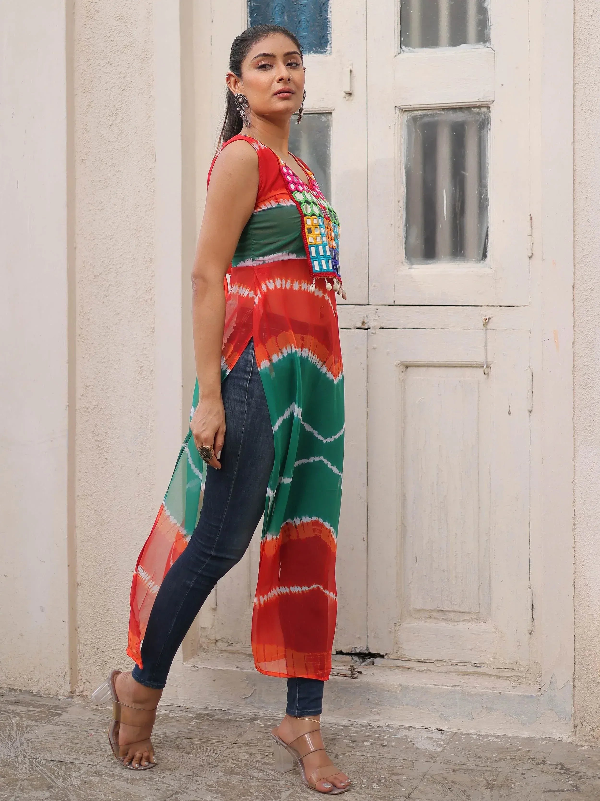 Woman in colorful tie-dye ethnic kurti with mirror work, paired with jeans and heels, standing by white door
