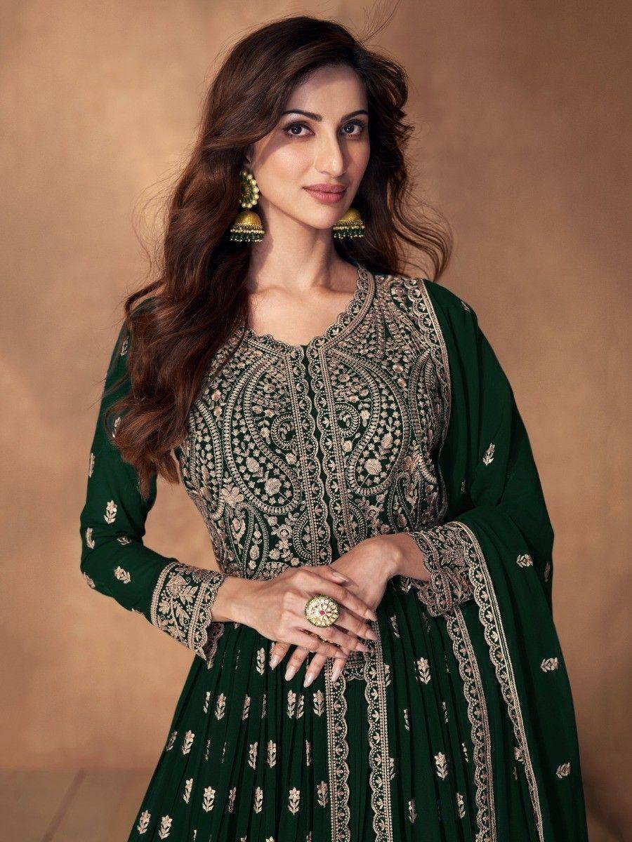 Woman wearing dark green embroidered ethnic gown with gold jewelry, posing indoors