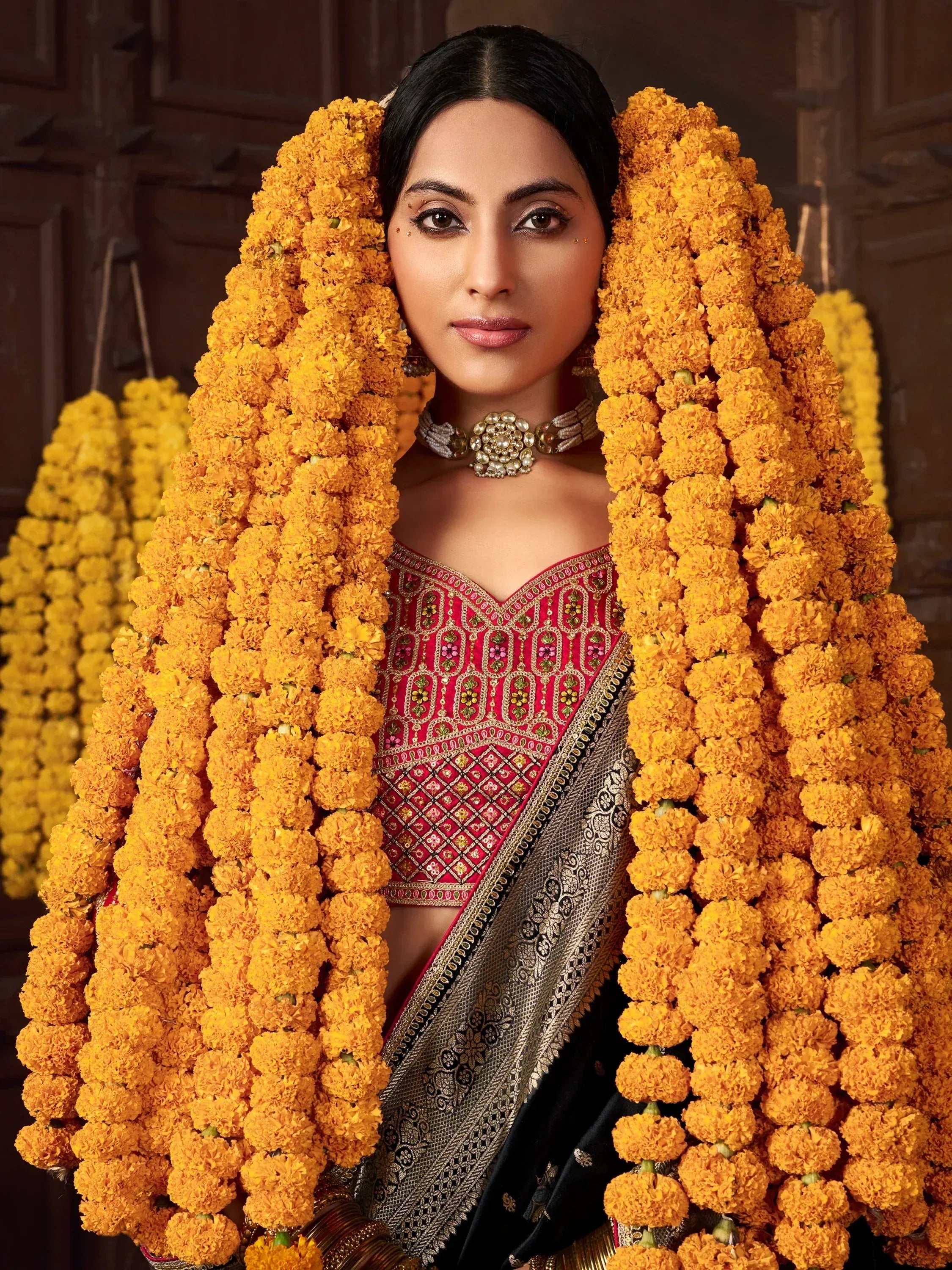Woman in red embroidered blouse and black saree with gold border, framed by marigold garlands, traditional Indian ethnic wear